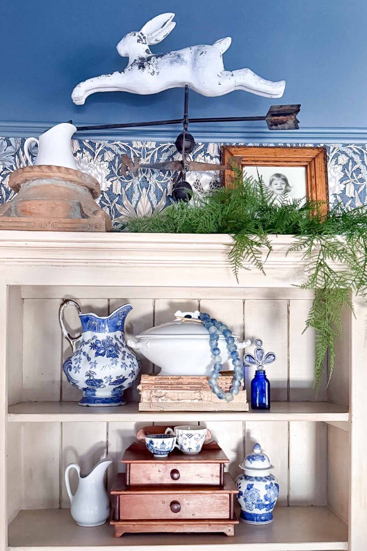 A shelf decorated with blue and white ceramics, a small pitcher, wooden boxes, a beaded necklace, green fern leaves, a framed photo, and a white rabbit figure leaping above the top shelf against a blue patterned wall.