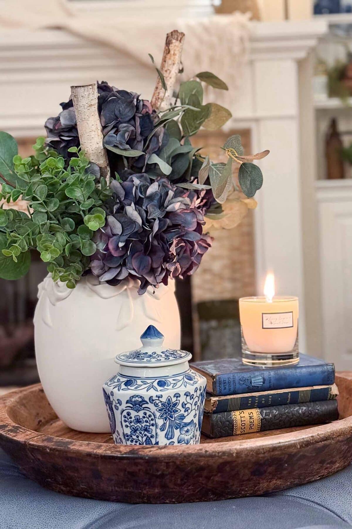 A wooden tray holds a white vase with purple and green flowers, a lit candle, a small blue and white jar, and a stack of three vintage books, all arranged in a cozy home setting.