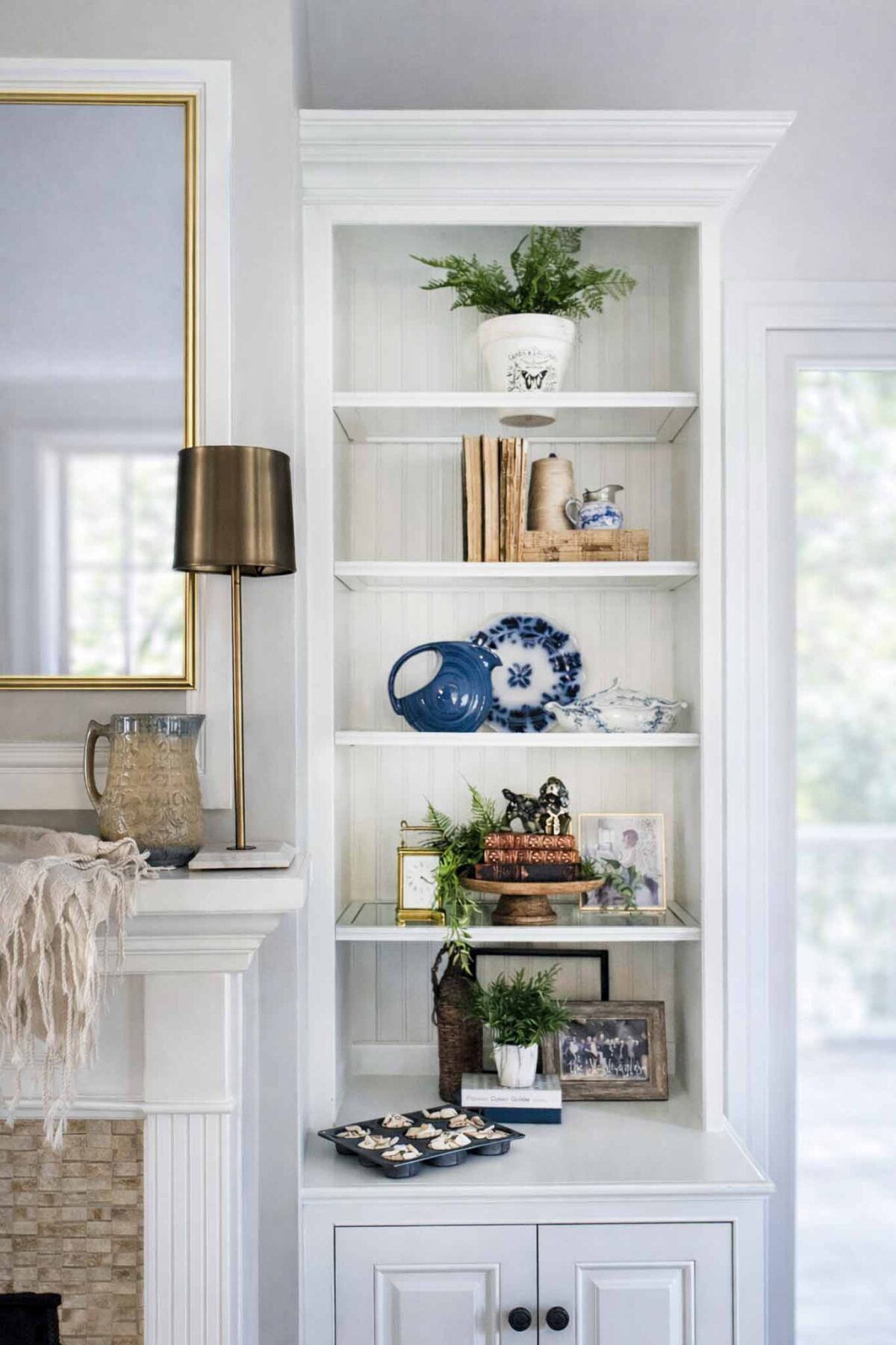 A white built-in shelf displays potted plants, vintage books, blue and white dishes, framed photos, and decorative objects beside a fireplace with a lamp and pitcher on the mantel.