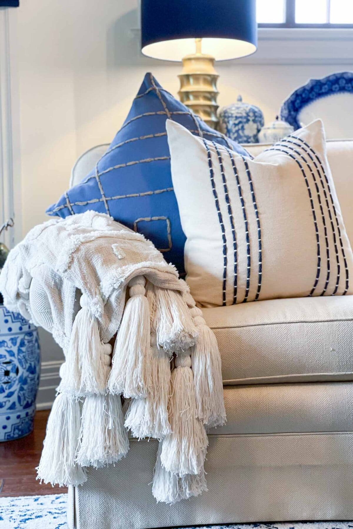 A beige sofa with two decorative pillows, one blue with stitched lines and one cream with vertical blue stripes, sits beside a cream throw blanket with large tassels. A blue and white lamp and vase are in the background.