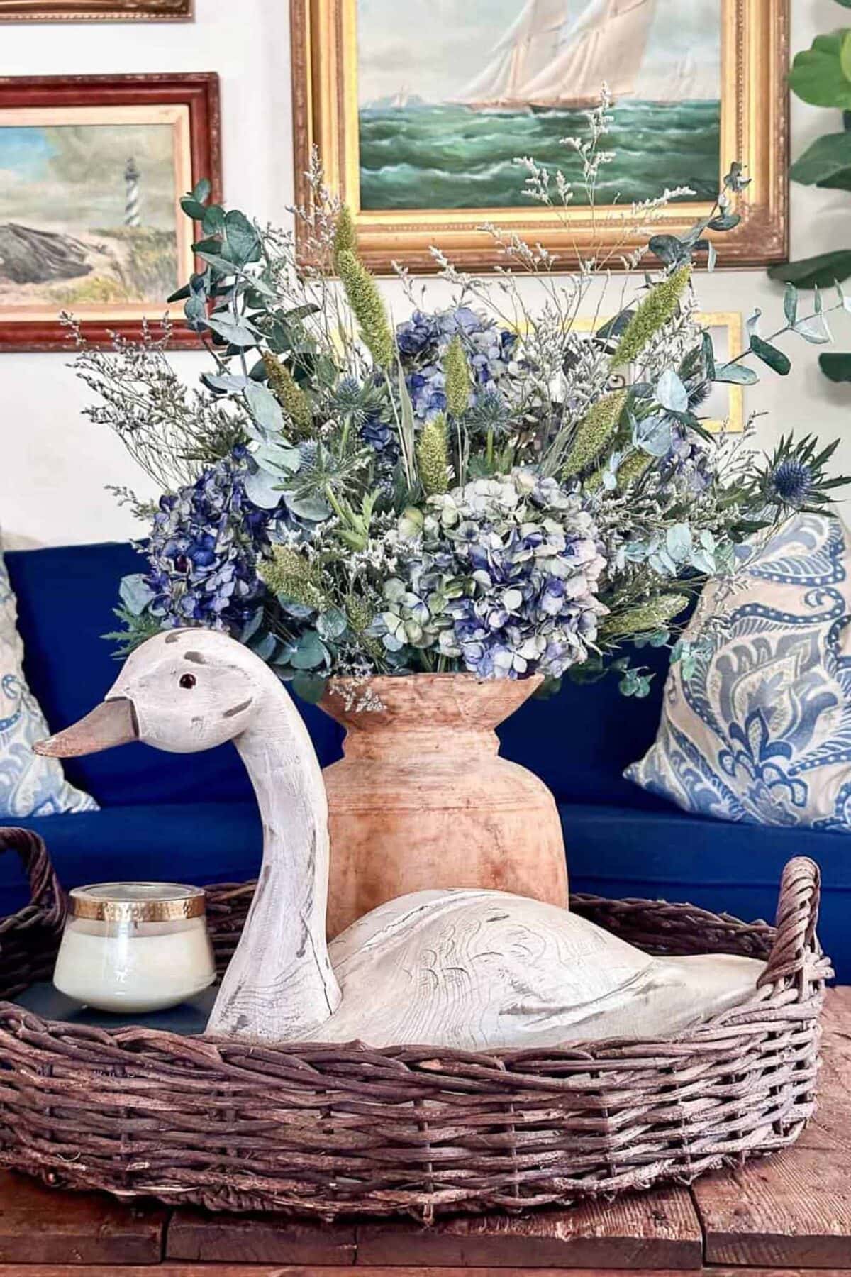 A wicker tray holds a white wooden swan figurine and a candle, set on a table in front of a bouquet of blue and white flowers in a clay vase, with framed nautical artwork and patterned pillows in the background.