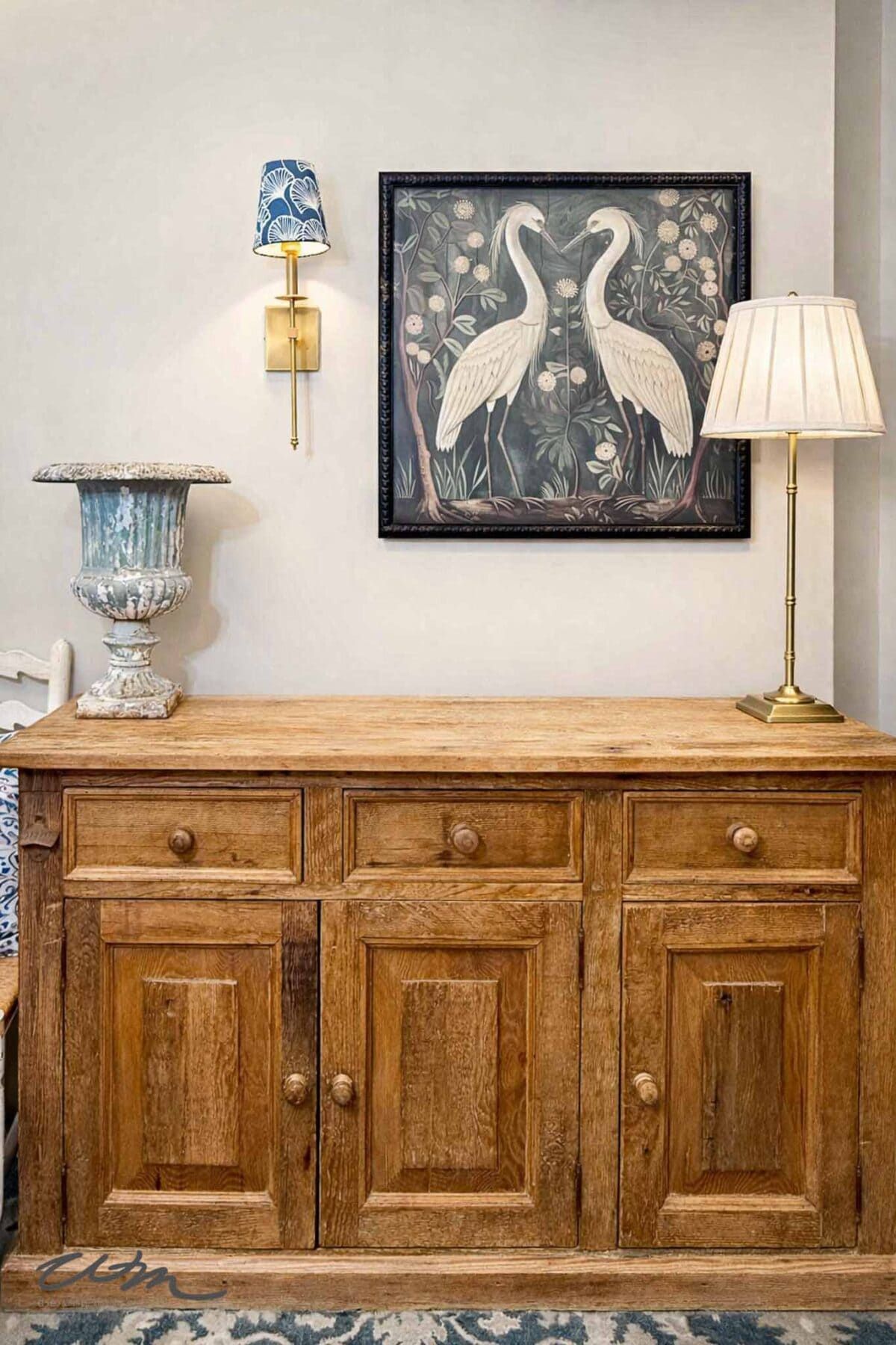 A rustic wooden sideboard brings vintage charm to the space, sitting below framed heron artwork. A brass table lamp, decorative blue vase, and a wall sconce with a blue patterned shade create perfect spring entry table decor.