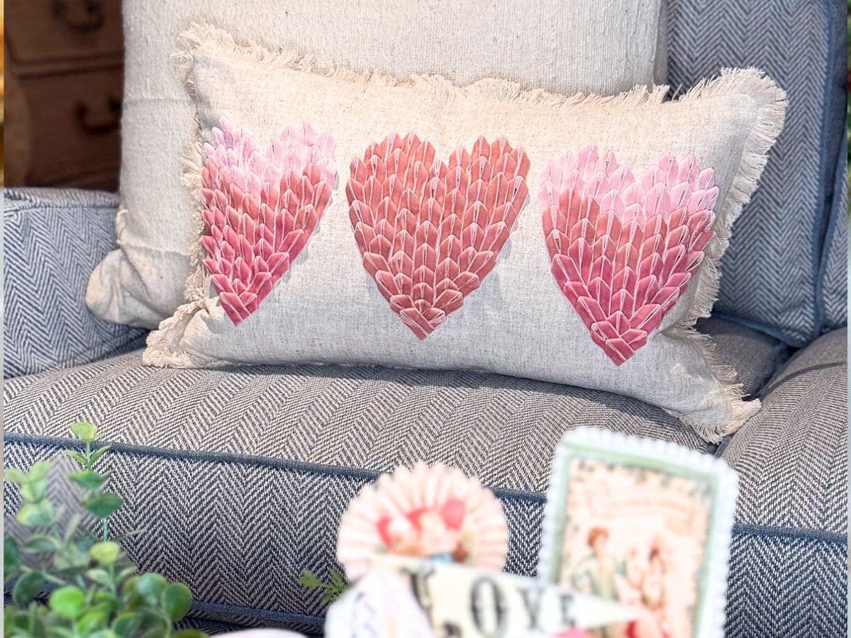A rectangular pillow with three pink and red heart designs, reminiscent of heart sugar cookies, sits on a blue and white herringbone-patterned armchair. Decorative items and greenery are visible in the foreground.