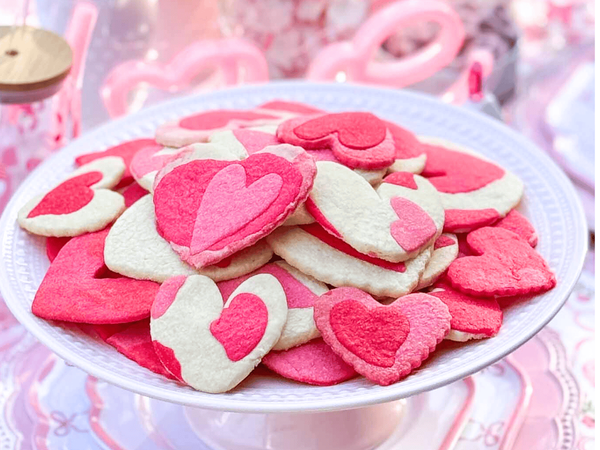 A white cake stand holds Heart Sugar Cookies in shades of pink, red, and white, each decorated with layered heart designs. The background features soft pink and white décor, creating a festive, romantic atmosphere.