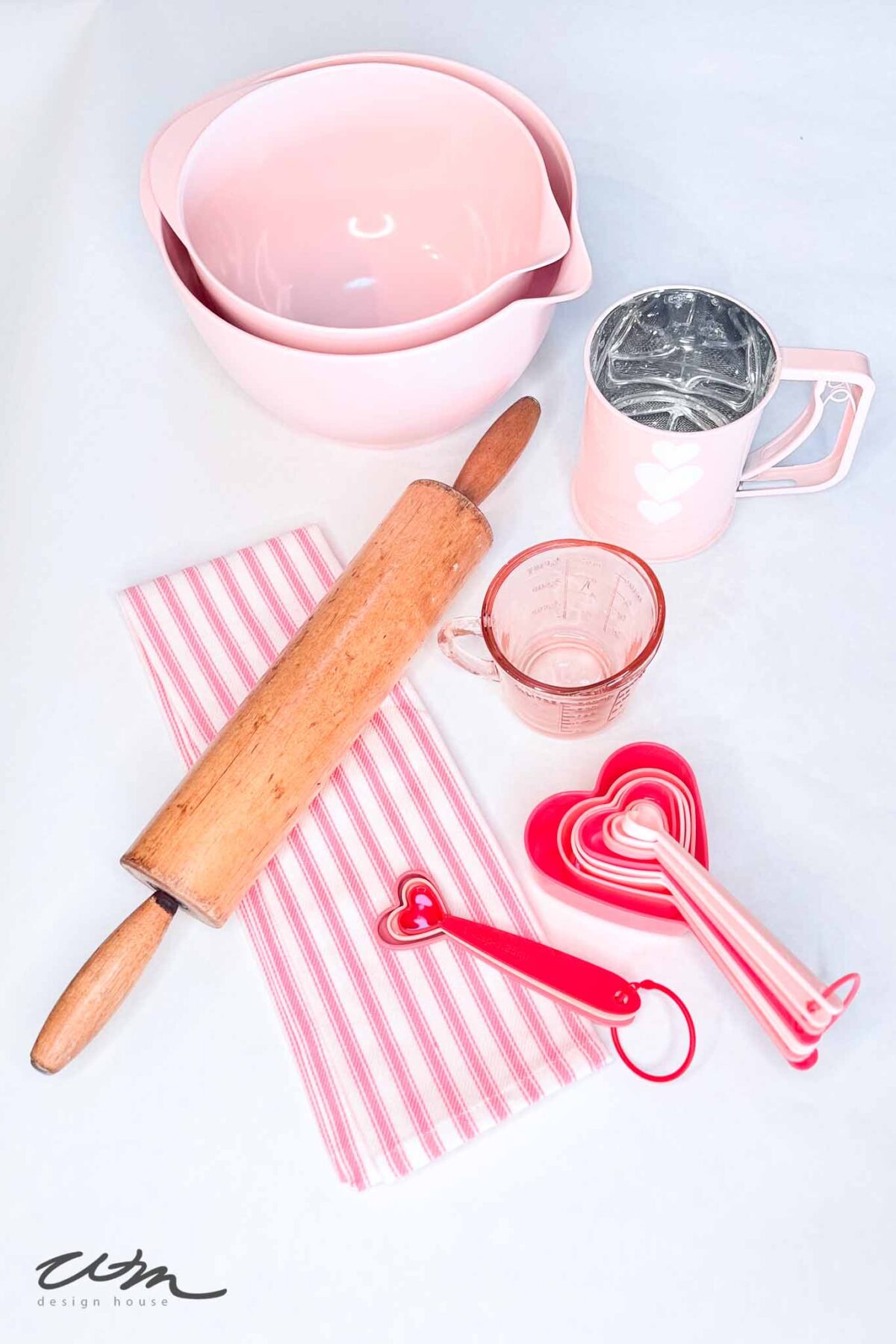 Baking utensils on a white surface, including a rolling pin, two pink mixing bowls, a pink flour sifter, a glass measuring cup, striped pink dish towel, and pink heart-shaped measuring spoons and cups.