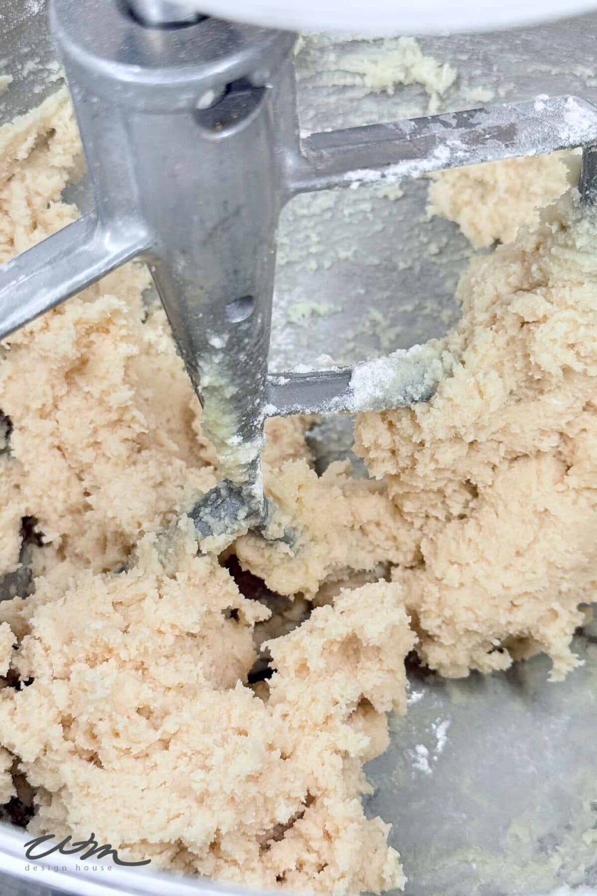 Close-up of cookie dough being mixed in a stand mixer with a metal paddle attachment. The dough appears light and crumbly, partially sticking to the sides and blade.