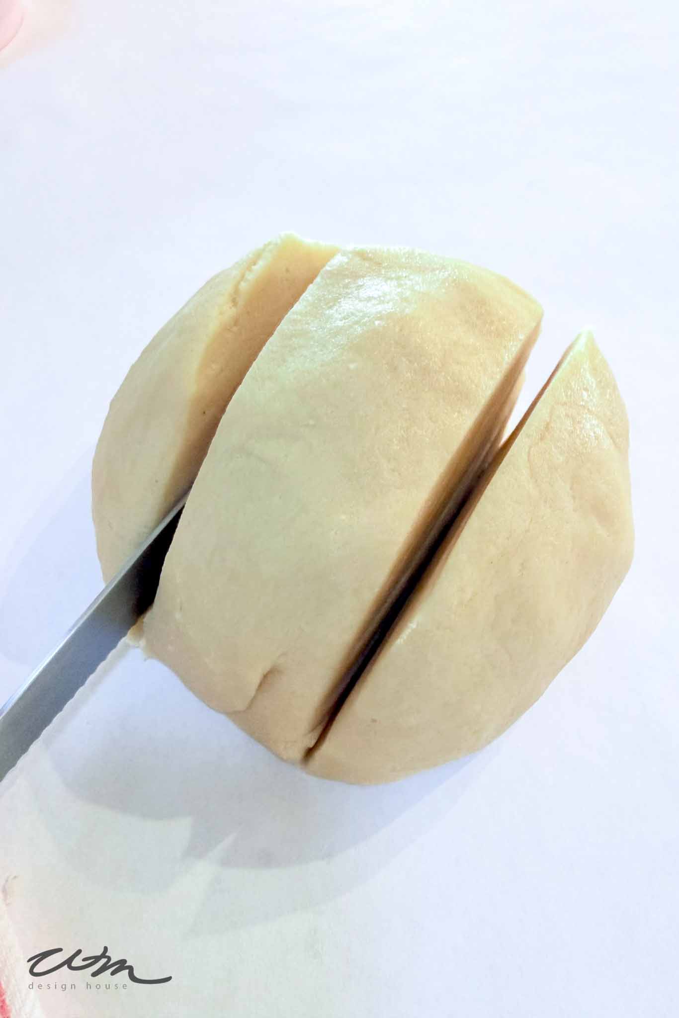 A round piece of dough being sliced into three sections with a serrated knife on a white surface.