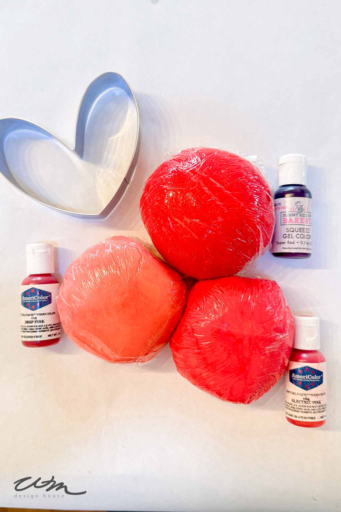 Three balls of red and pink dough wrapped in plastic, four bottles of food coloring, and a metal heart-shaped cookie cutter arranged on a white surface.