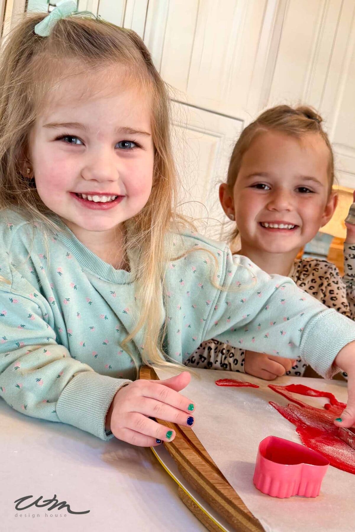Two young children smile while making cookies. The girl in front, wearing a light green sweater, presses dough with a red cookie cutter, while the other child in a spotted shirt stands behind her. Both look happy in a kitchen setting.