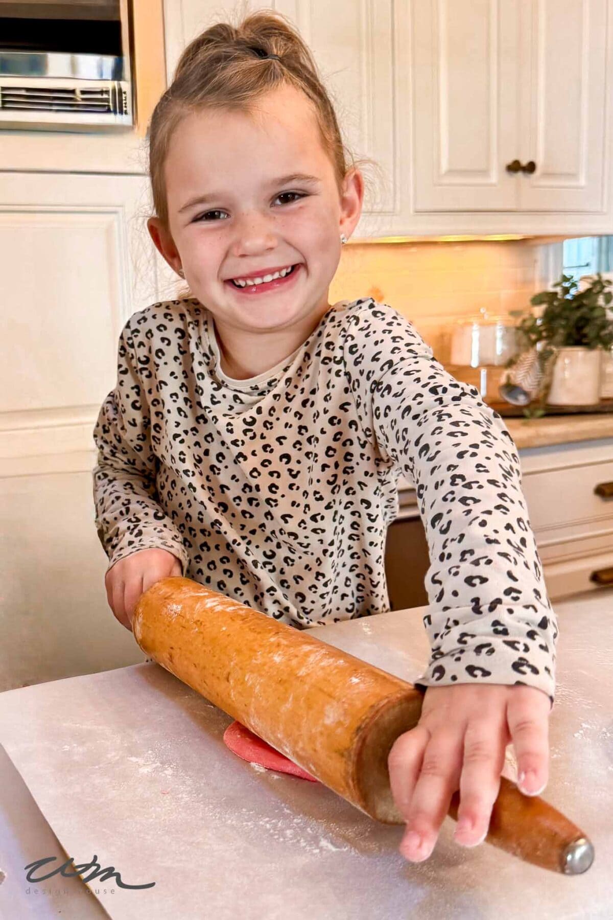 Smiling young girl in a leopard-print top rolling out dough with a wooden rolling pin on a floured surface in a bright kitchen.