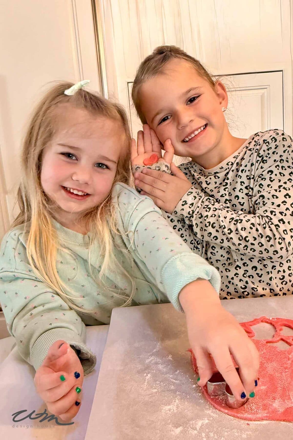 Two young girls smile at the camera while making cookies. One girl presses a cookie cutter into red dough, while the other holds up a small, heart-shaped dough cutout. Both are wearing long-sleeve shirts with cute prints.