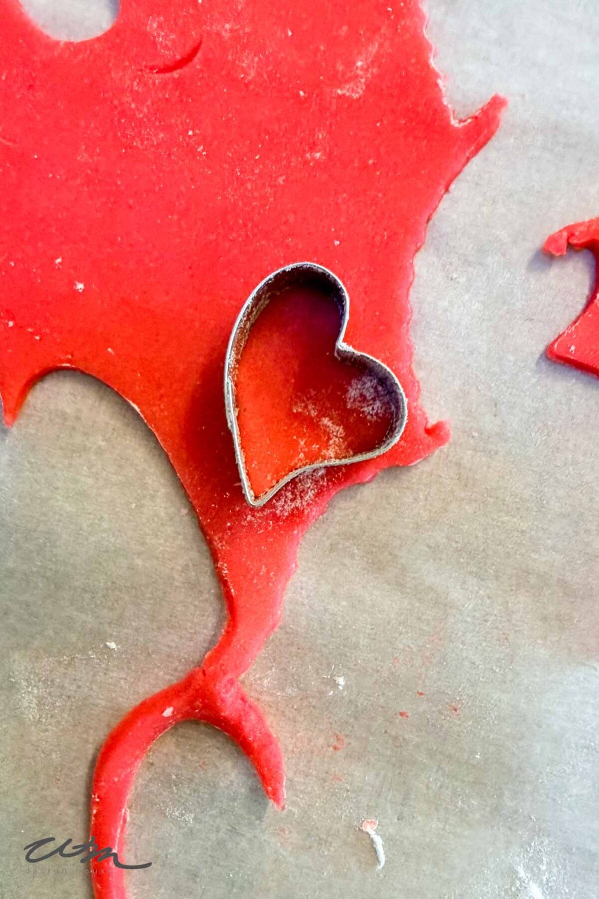 A heart-shaped metal cookie cutter sits on rolled-out red dough, partially cutting out a heart shape, with scattered dough scraps on a parchment-lined surface.