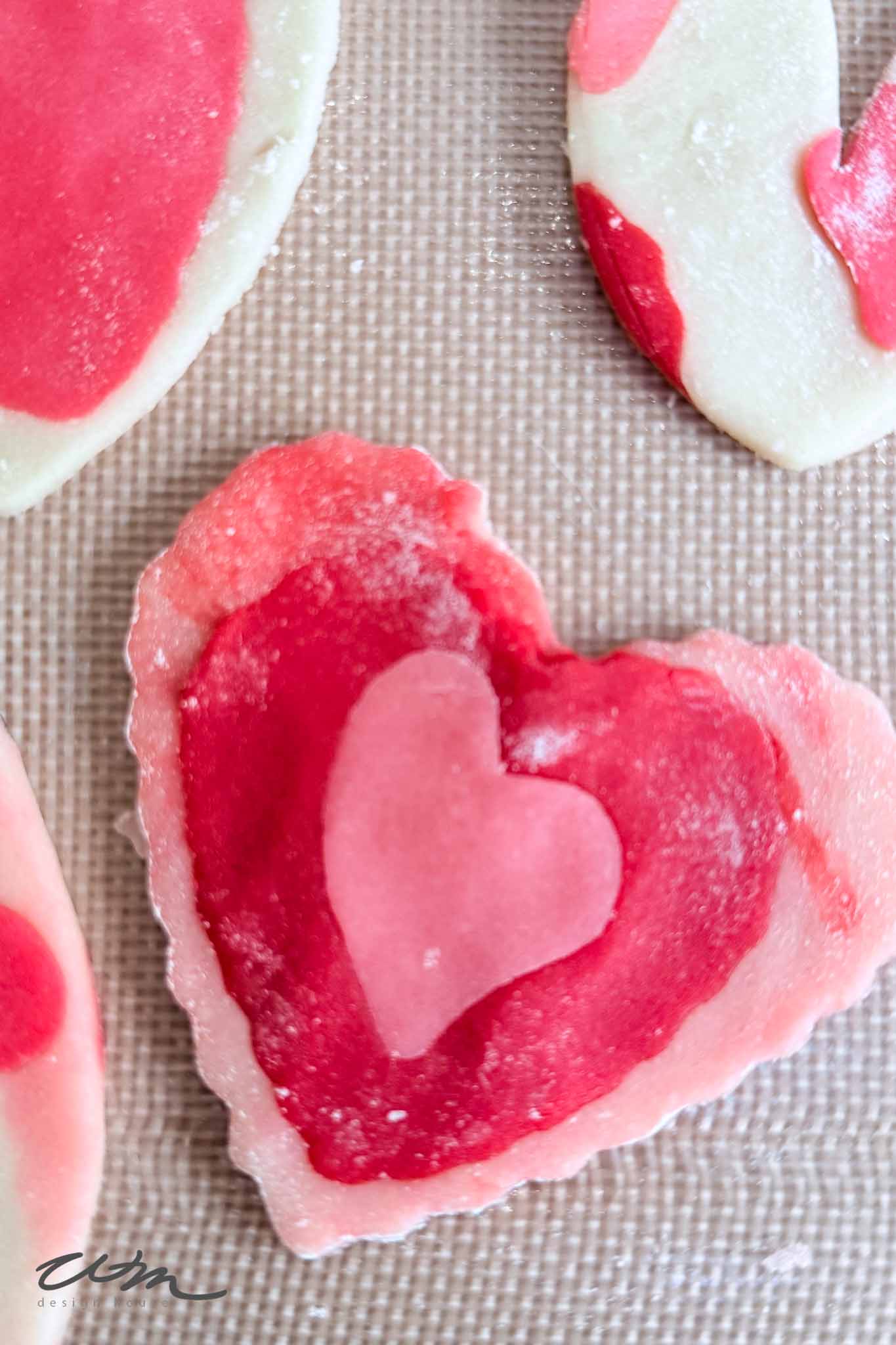 A heart-shaped cookie with pink and red marbled patterns and a small pink heart in the center, placed on a textured baking mat alongside other colorful cookies.