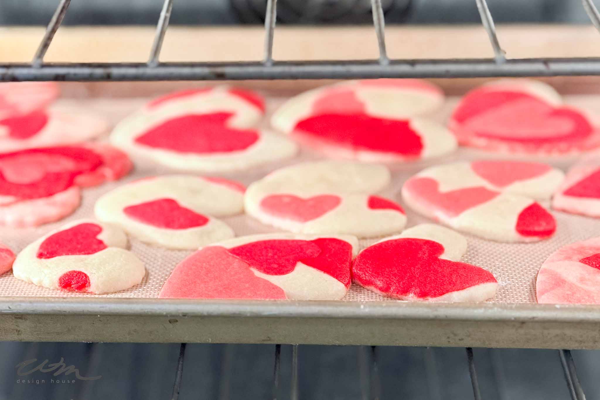 Heart-shaped pink, red, and white cookies are baking on a tray inside an oven. The cookies have a marbled, multi-color pattern and some are round while others are heart-shaped.