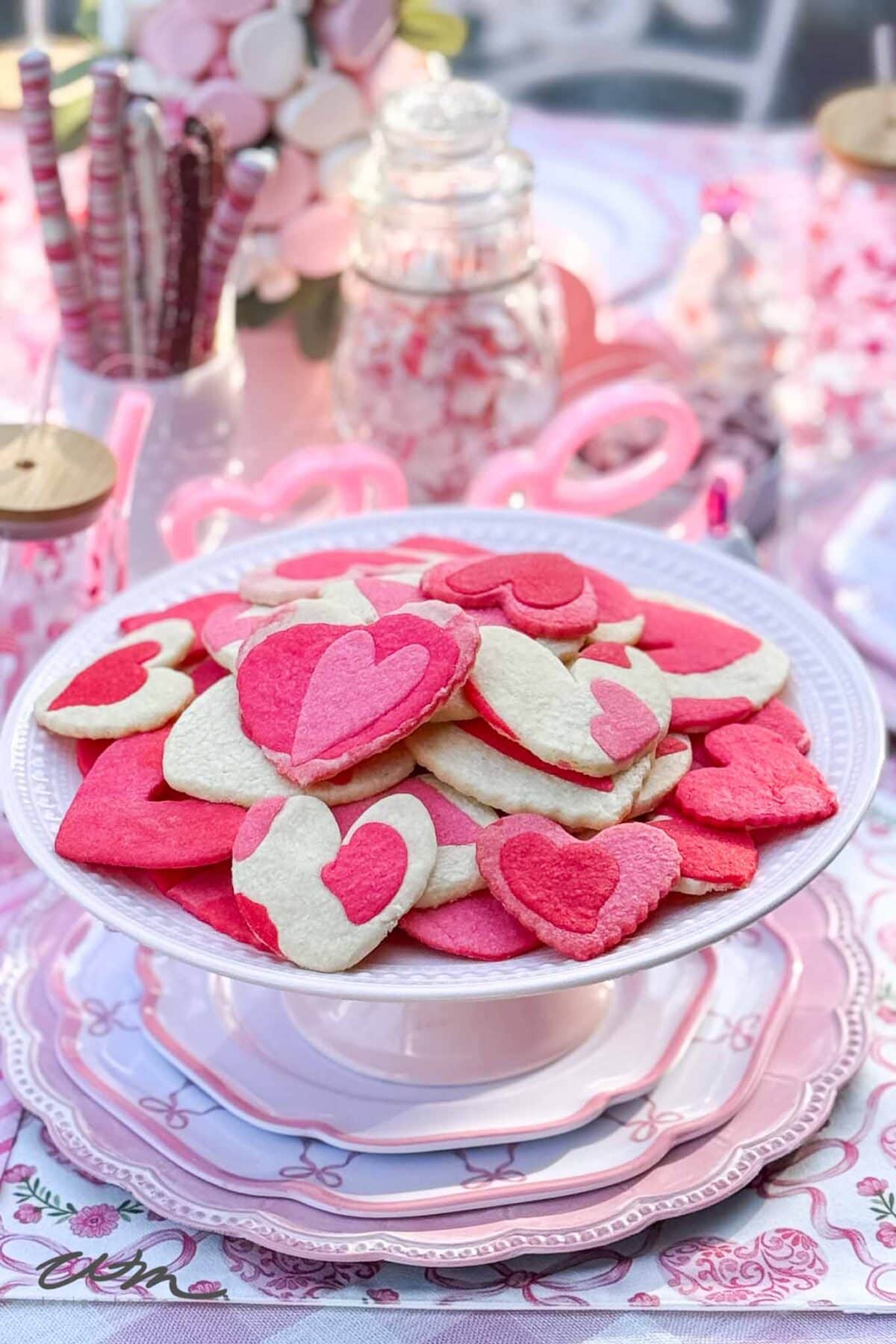 A white cake stand displays heart-shaped pink and white cookies, arranged on stacked pink and white plates. The table is decorated with other pink treats and heart-themed items, creating a festive, romantic atmosphere.