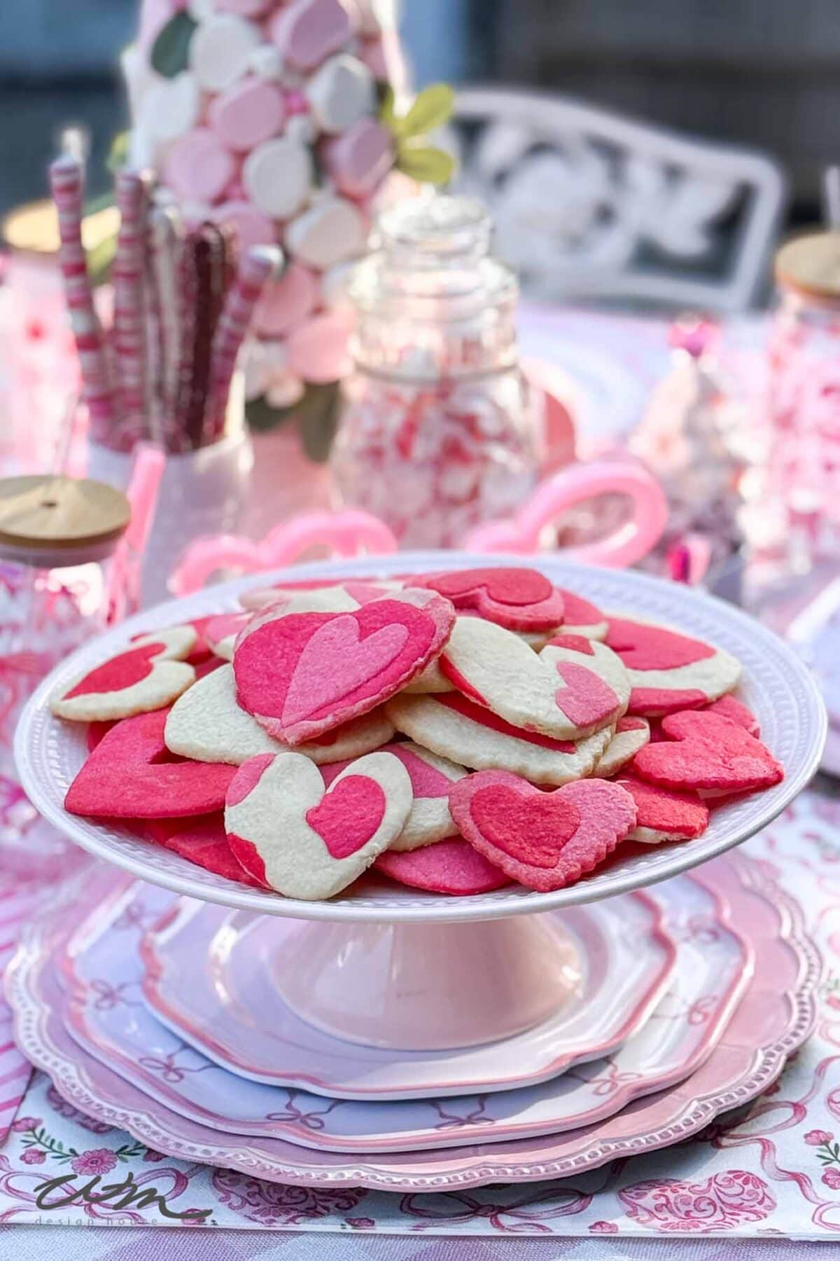A white cake stand holds heart-shaped pink and white cookies on a pastel table set with pink plates, glass jars, and festive Valentine’s Day decorations in the background.