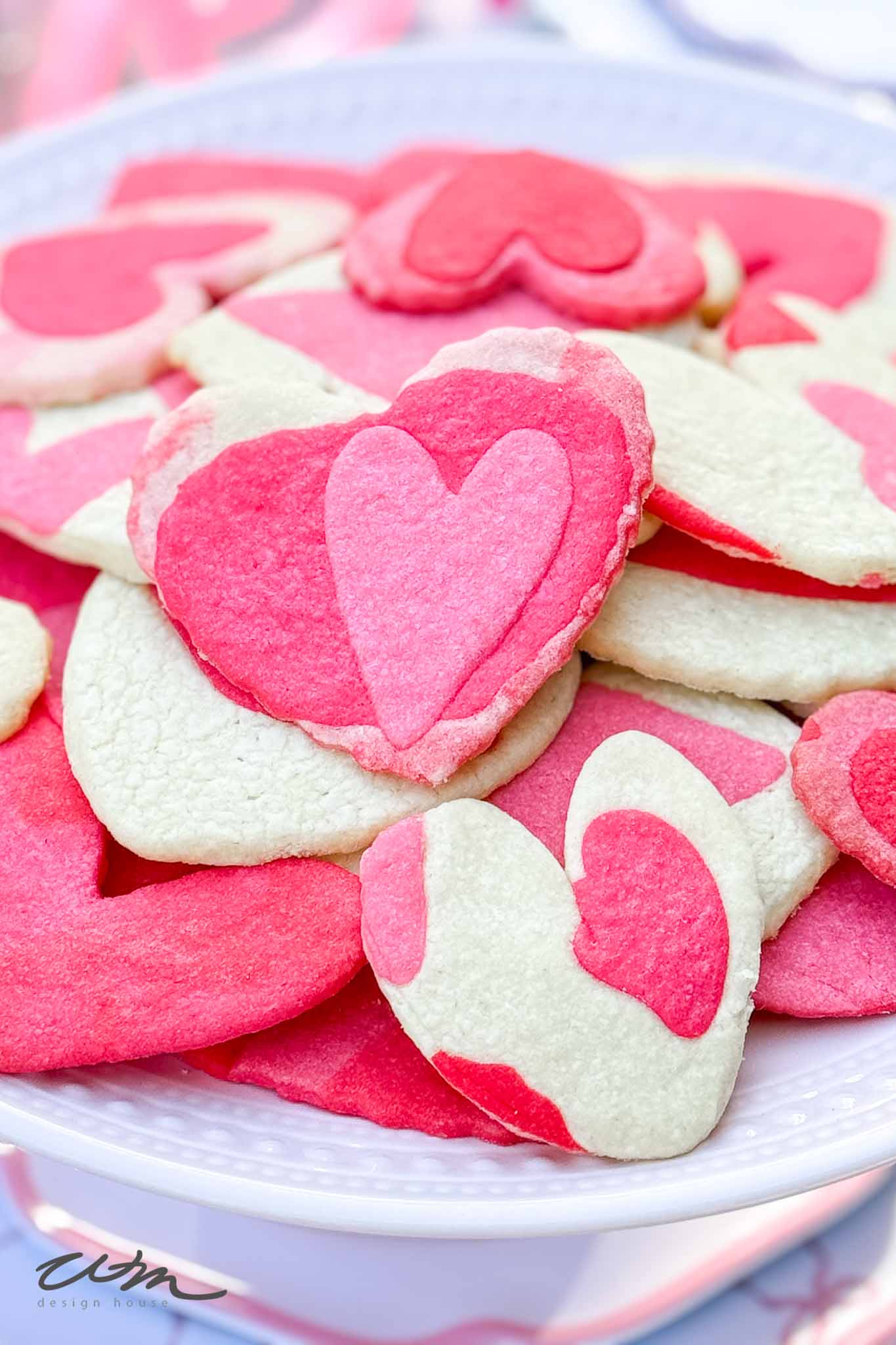A plate of heart-shaped sugar cookies in pink, red, and white colors, some decorated with smaller heart patterns on top. The cookies are arranged in a pile on a white plate.