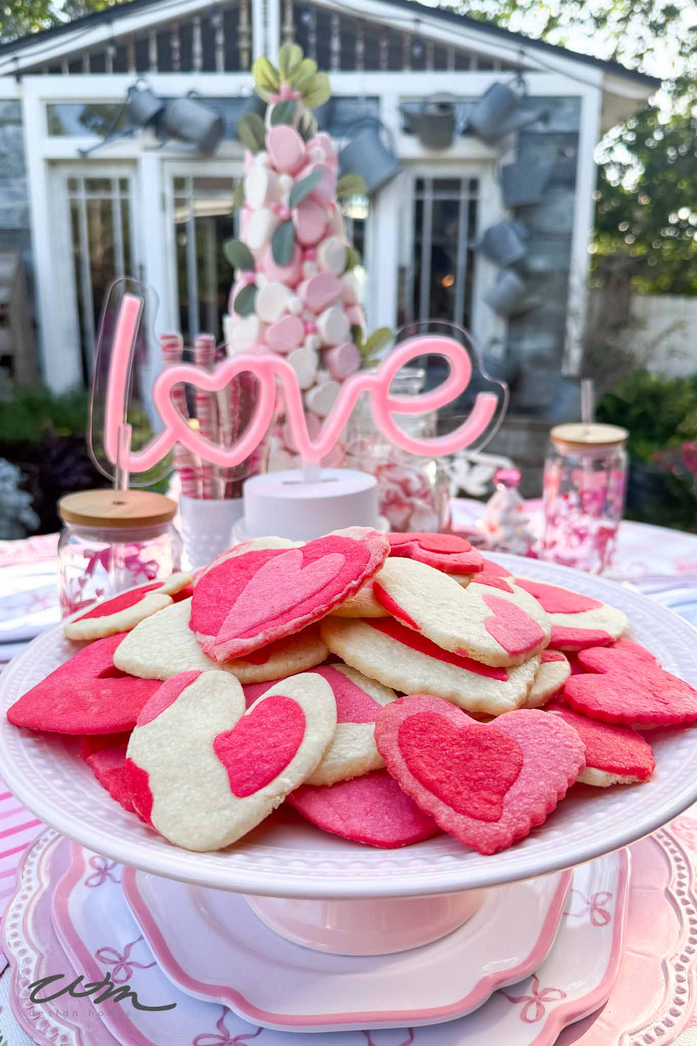 A plate of pink and white heart-shaped cookies sits on a table decorated for Valentine's Day, with a pink "love" sign and jars in the background. A garden shed is visible in the distance.
