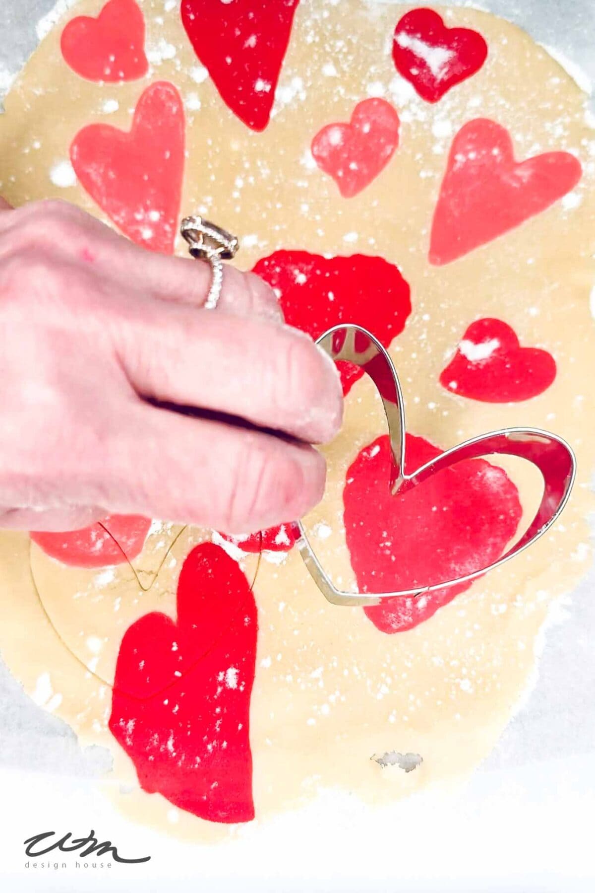 A hand holding a heart-shaped cookie cutter presses into rolled out dough decorated with red heart shapes, preparing to cut out cookies.