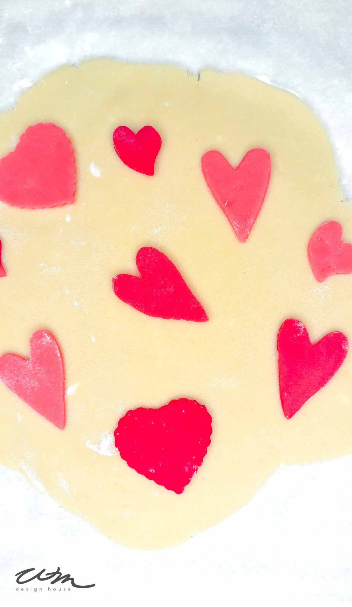 Heart Sugar Cookies: Heart-shaped red and pink cutouts placed on rolled-out cookie dough, ready for baking, on a white surface.