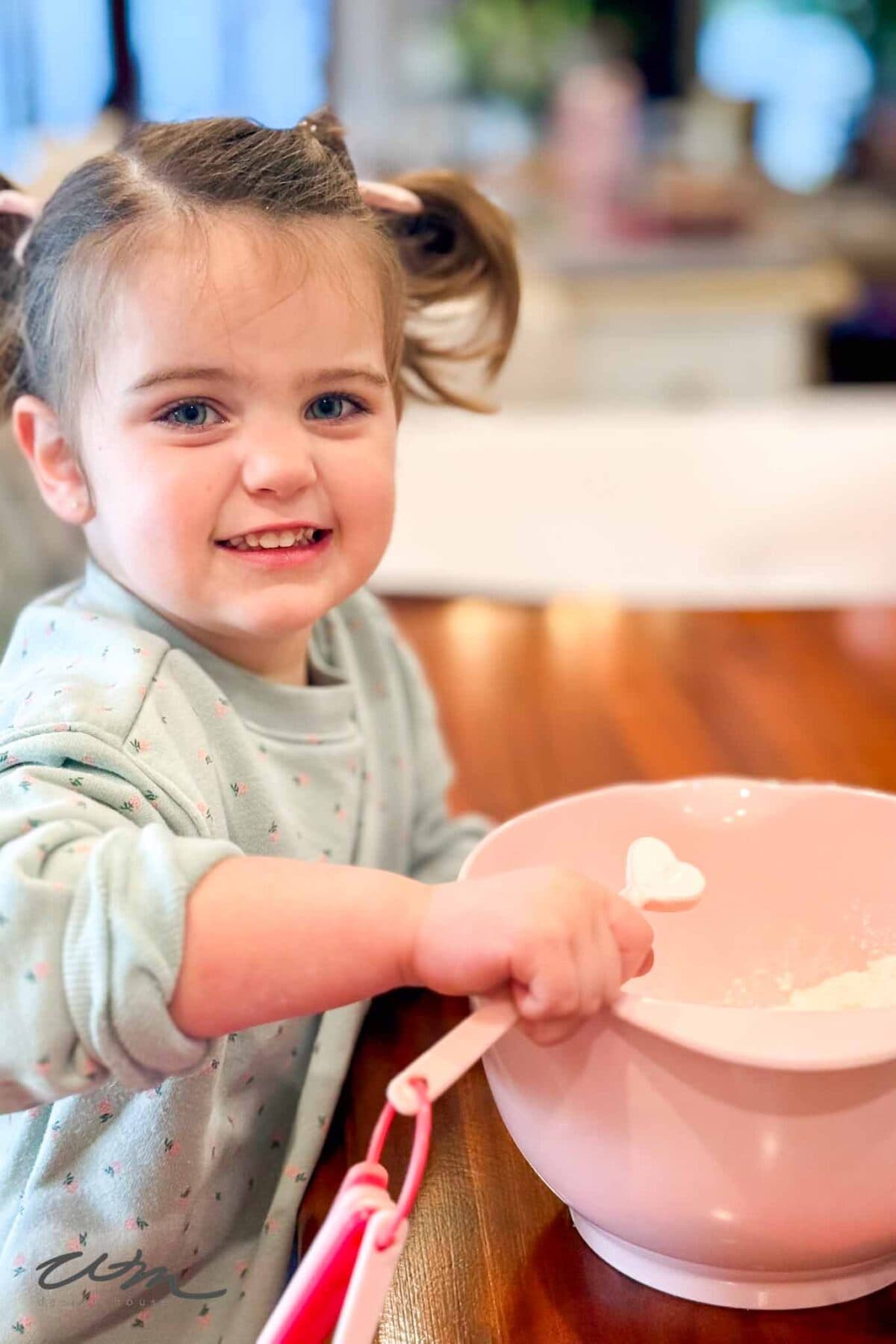 A young girl with pigtails smiles while stirring ingredients for Heart Sugar Cookies in a pink mixing bowl with a white spoon, sitting at a wooden table in a bright, cozy kitchen.