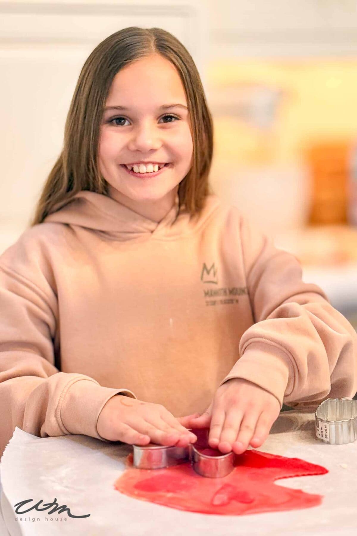 A smiling girl in a beige hoodie uses a cookie cutter on red dough placed on parchment paper, preparing heart sugar cookies in a bright kitchen.