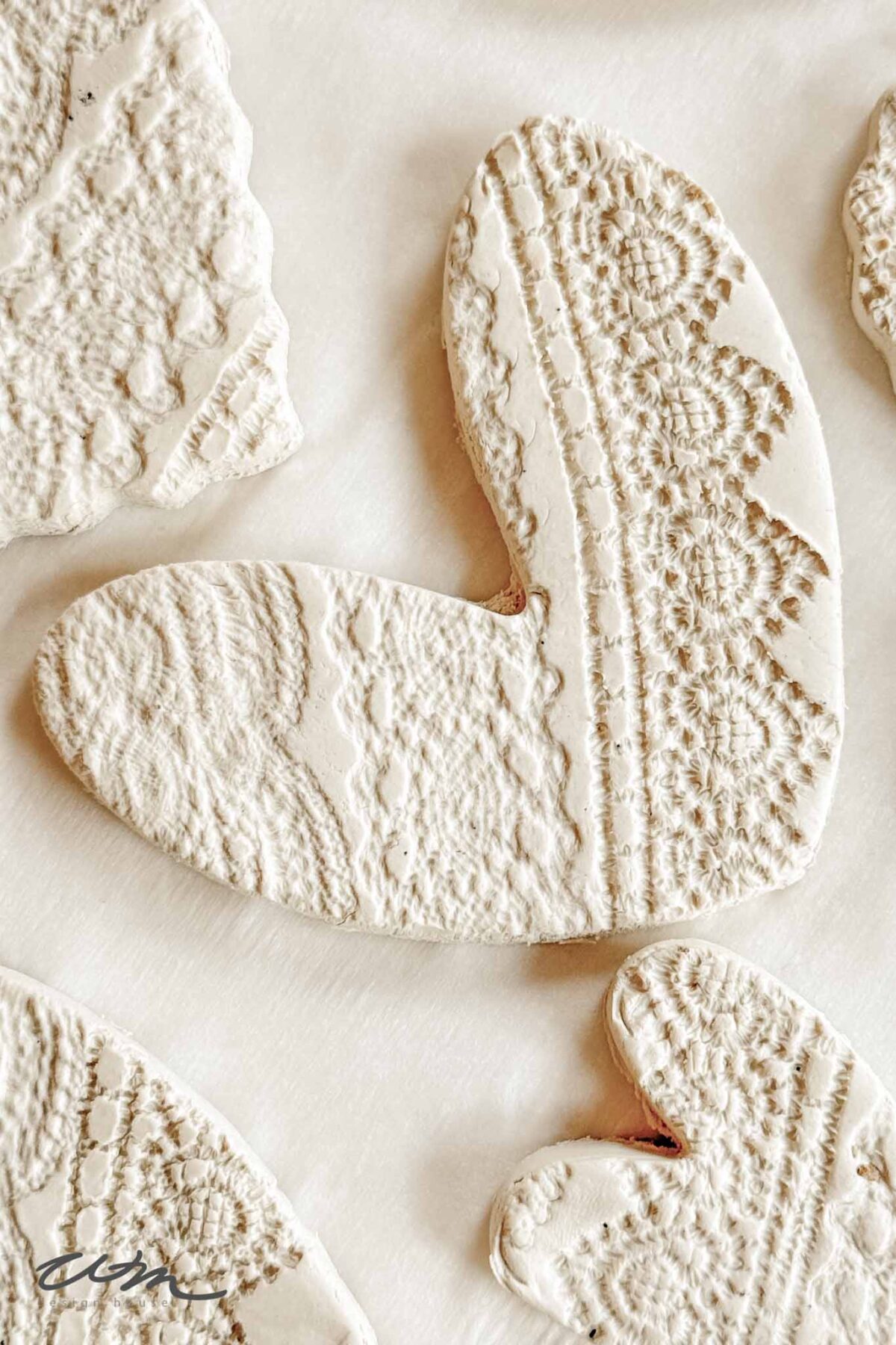 A white clay ornament shaped like a heart with intricate lace texture pressed into the surface, lying on a light background. Other similarly textured clay shapes are partially visible around it.