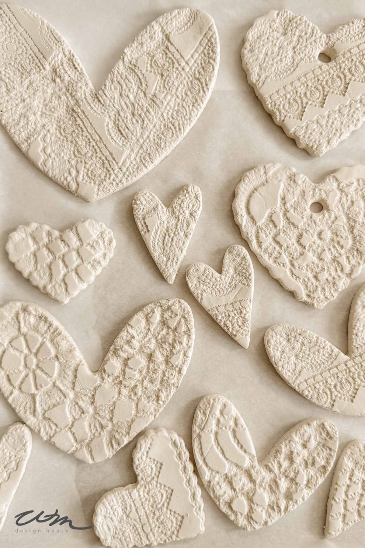 A collection of textured, cream-colored clay hearts with intricate lace patterns, arranged on a light surface. Some hearts have holes, suggesting they could be used as ornaments or decorations.
