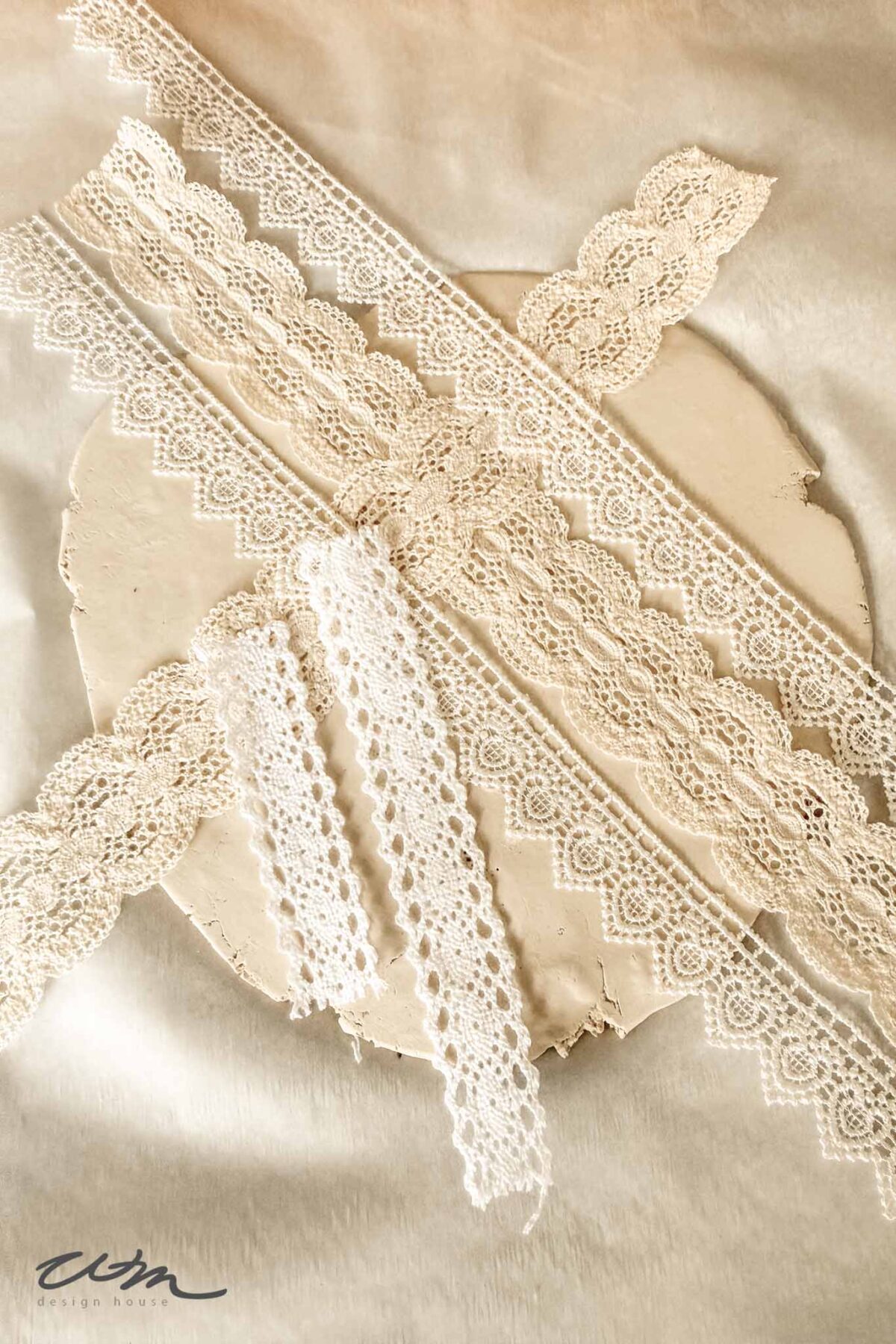 Several strips of delicate, cream and white lace are arranged on a round, textured beige surface set against a soft, neutral background. Different lace patterns and widths are displayed.