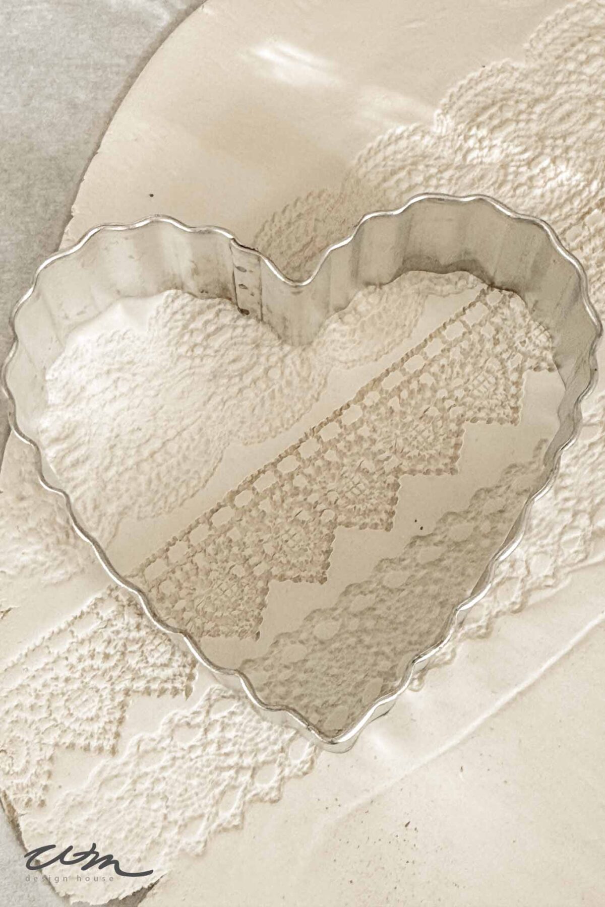 A scalloped metal heart-shaped cookie cutter rests on a textured white surface with lace patterns and soft lighting, creating a delicate, vintage look.