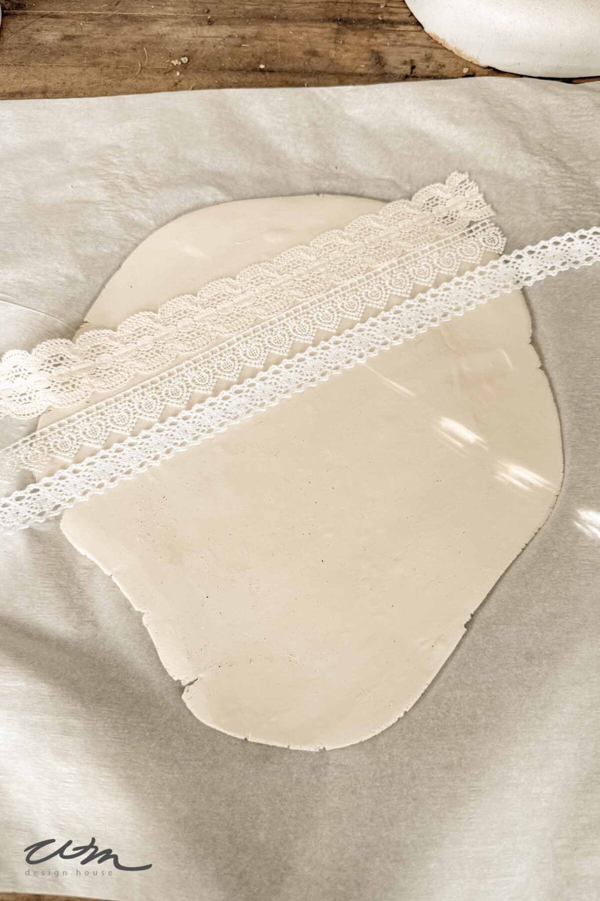 A piece of white clay rolled flat on parchment paper with two strips of white lace placed diagonally across it, ready for pressing to create a textured pattern.
