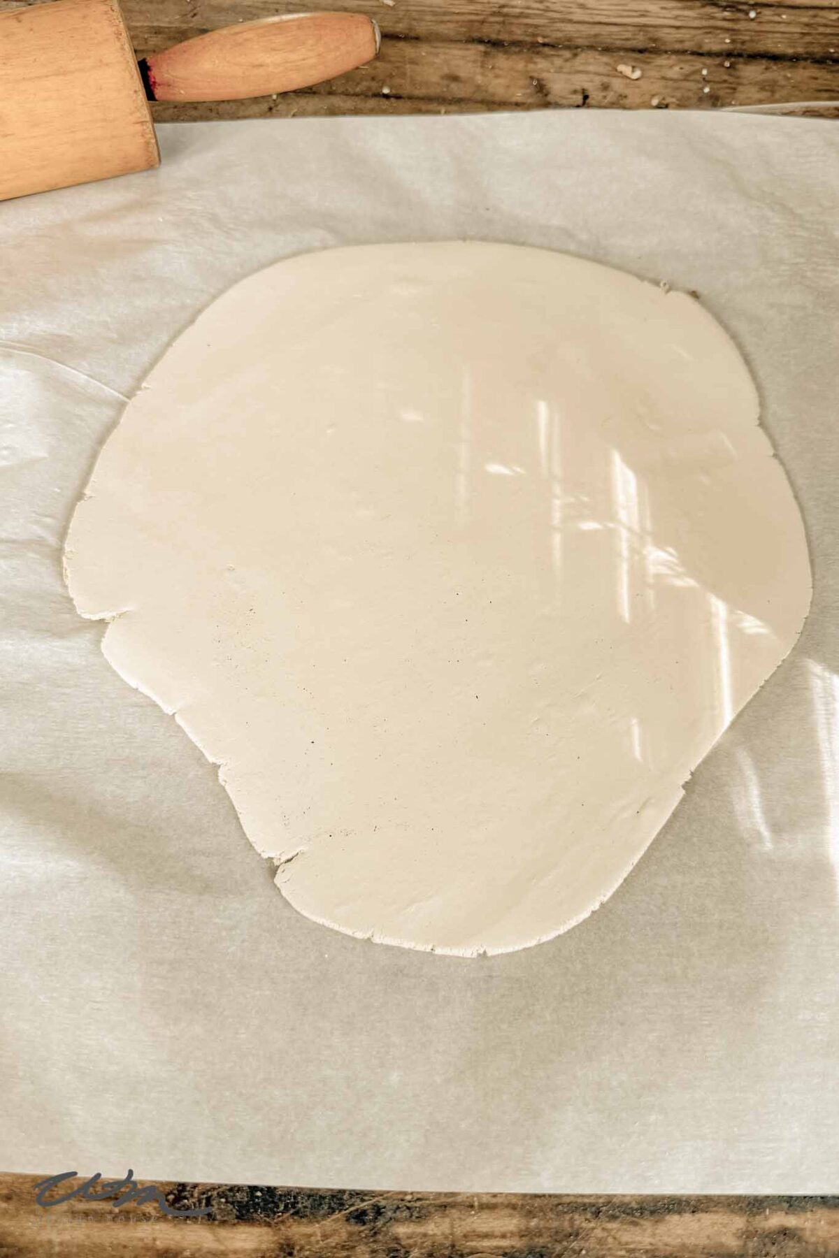 A sheet of white clay rolled out on parchment paper, with a wooden rolling pin partially visible at the top on a rustic wooden surface.