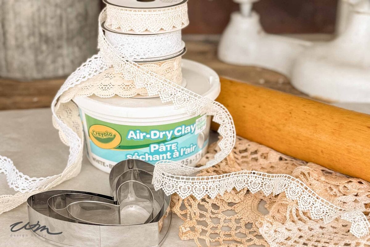 A tub of Crayola air-dry clay, lace ribbons, heart-shaped cookie cutters, a rolling pin, and a crocheted doily are arranged on a table, suggesting a vintage-inspired craft setup.