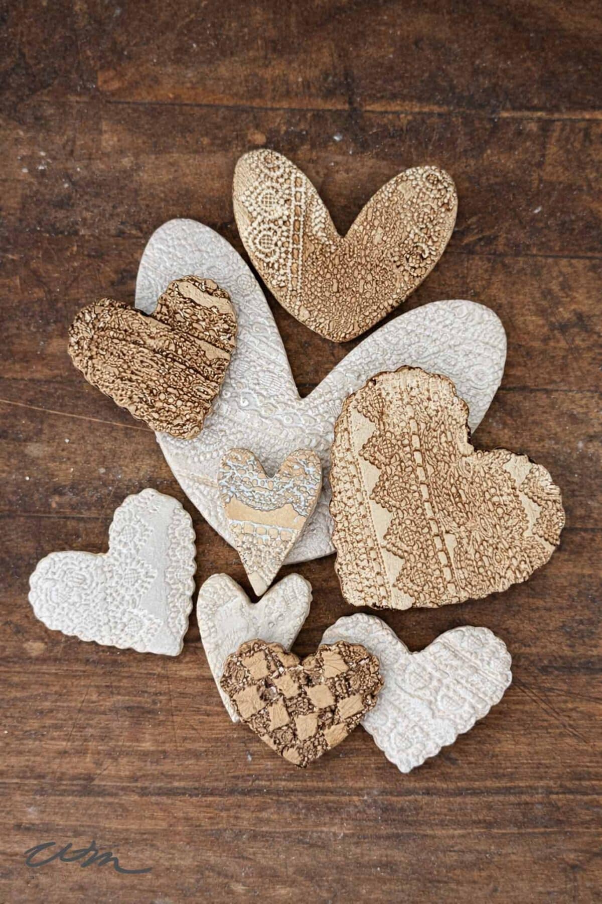Several textured lace hearts in shades of beige and brown are arranged on a wooden surface. The hearts feature intricate lace and patterned details, creating a rustic and handmade appearance.