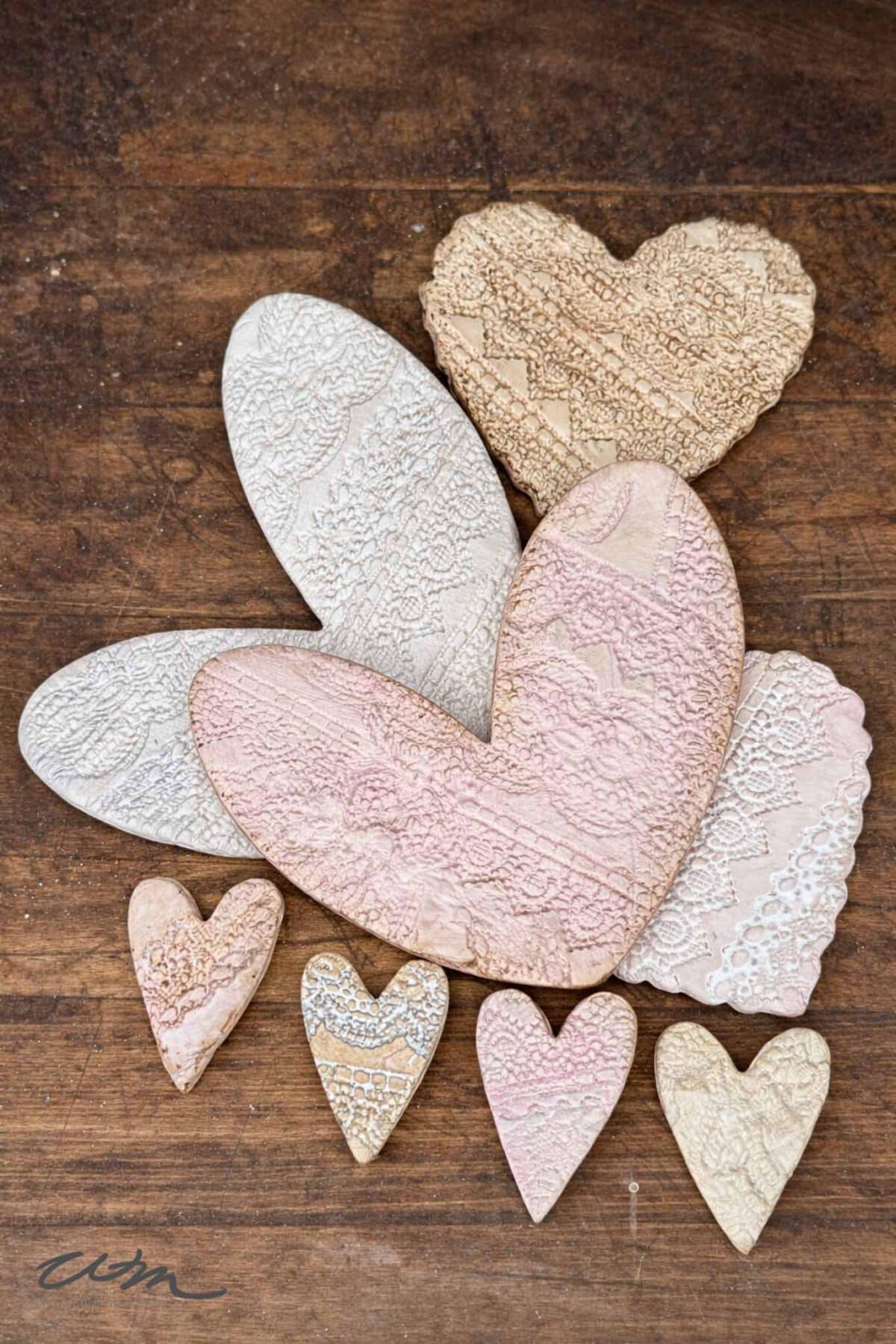 Several lace hearts, crafted into heart-shaped cookies with delicate patterns in pastel shades of pink, white, and beige, are arranged on a rustic wooden surface.