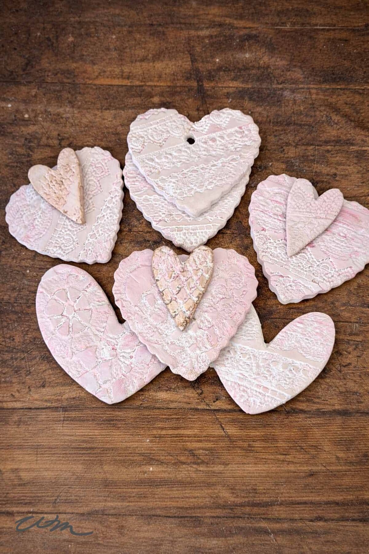 Six decorative Lace Hearts made from textured clay, painted in soft pink and white tones with intricate lace patterns, arranged on a wooden surface.