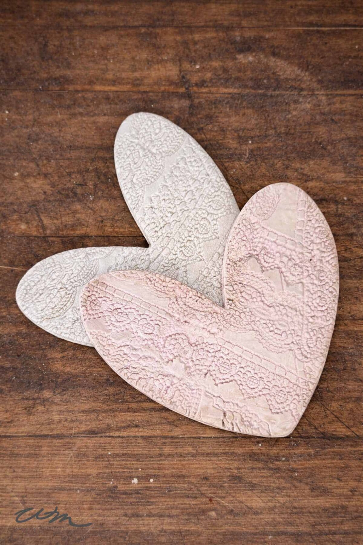 Two textured heart-shaped ceramic pieces, one light beige and one pale pink, are overlapping on a rustic wooden surface. Both Lace Hearts feature intricate lace-like patterns imprinted on them.