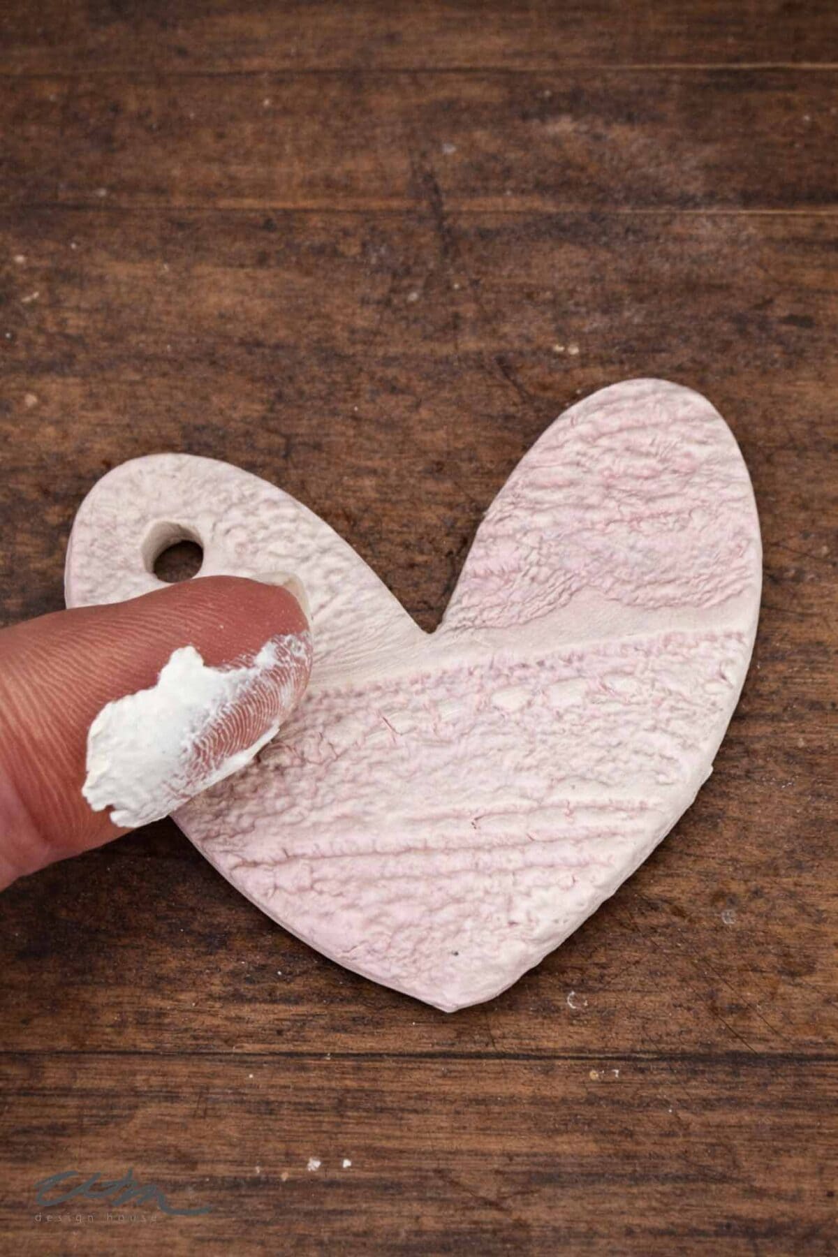 A finger with white paint touches a textured, heart-shaped clay pendant with a small hole, reminiscent of delicate Lace Hearts, resting on a wooden surface.