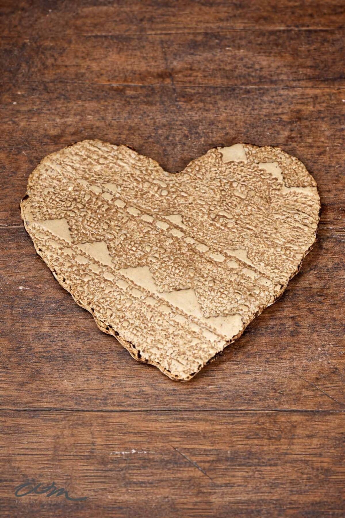 A heart-shaped piece of textured cardboard lies on a wooden surface. The cardboard, reminiscent of Lace Hearts, displays a corrugated pattern and rough edges.