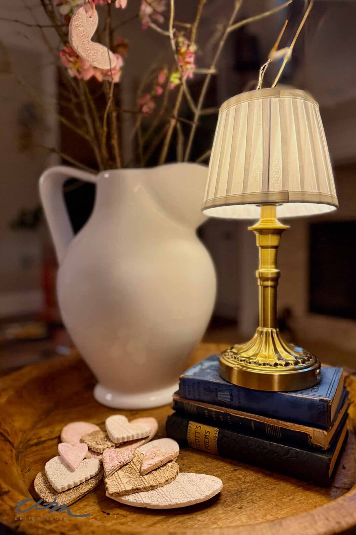 A small table lamp sits on a stack of vintage books next to a white ceramic pitcher with pink blossoms. Wooden and lace hearts with pink accents rest on a tray in the foreground, adding to the cozy, romantic atmosphere.