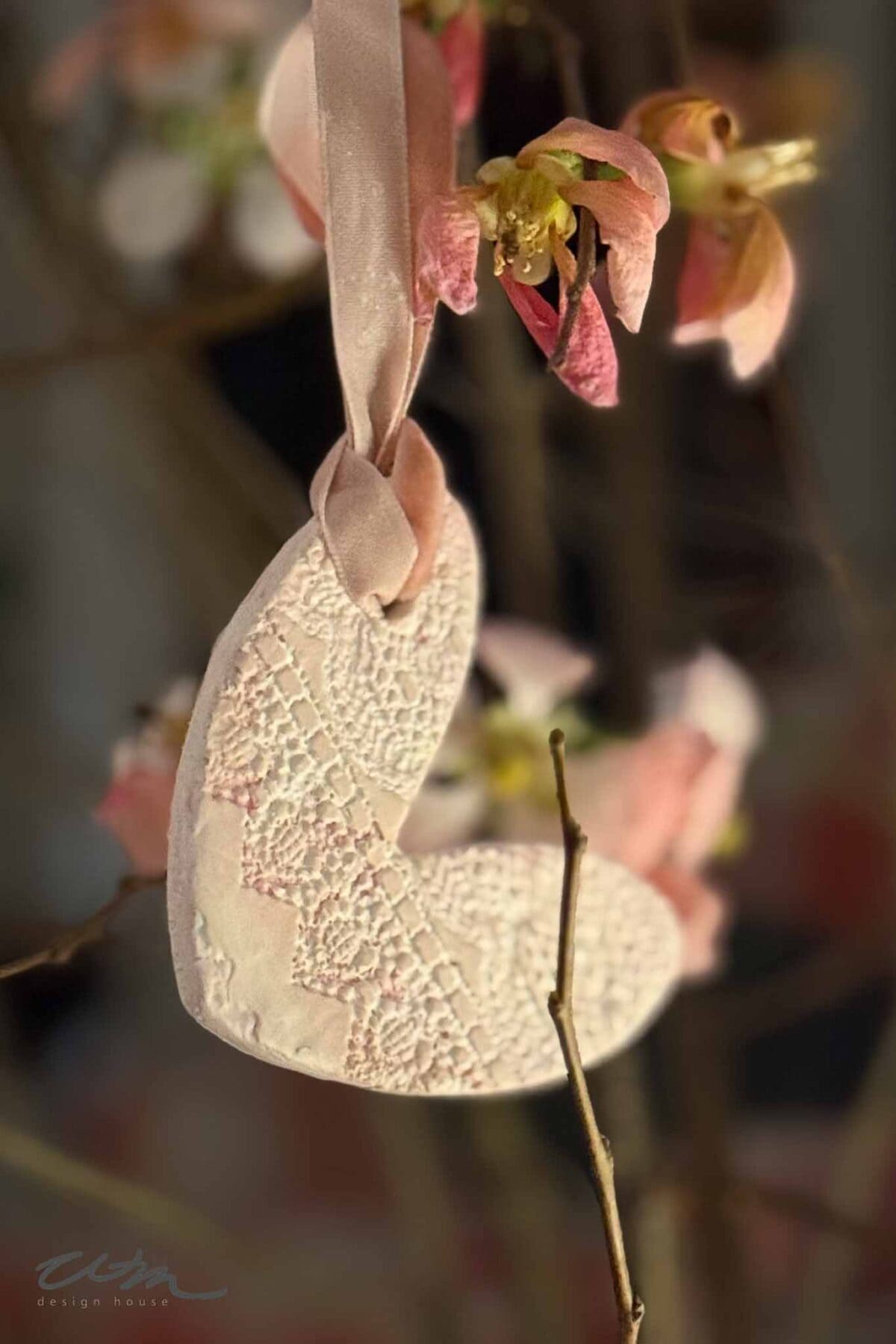 A textured ceramic heart ornament from the Lace Hearts collection with a pink ribbon hangs from a branch adorned with softly focused pink blossoms.
