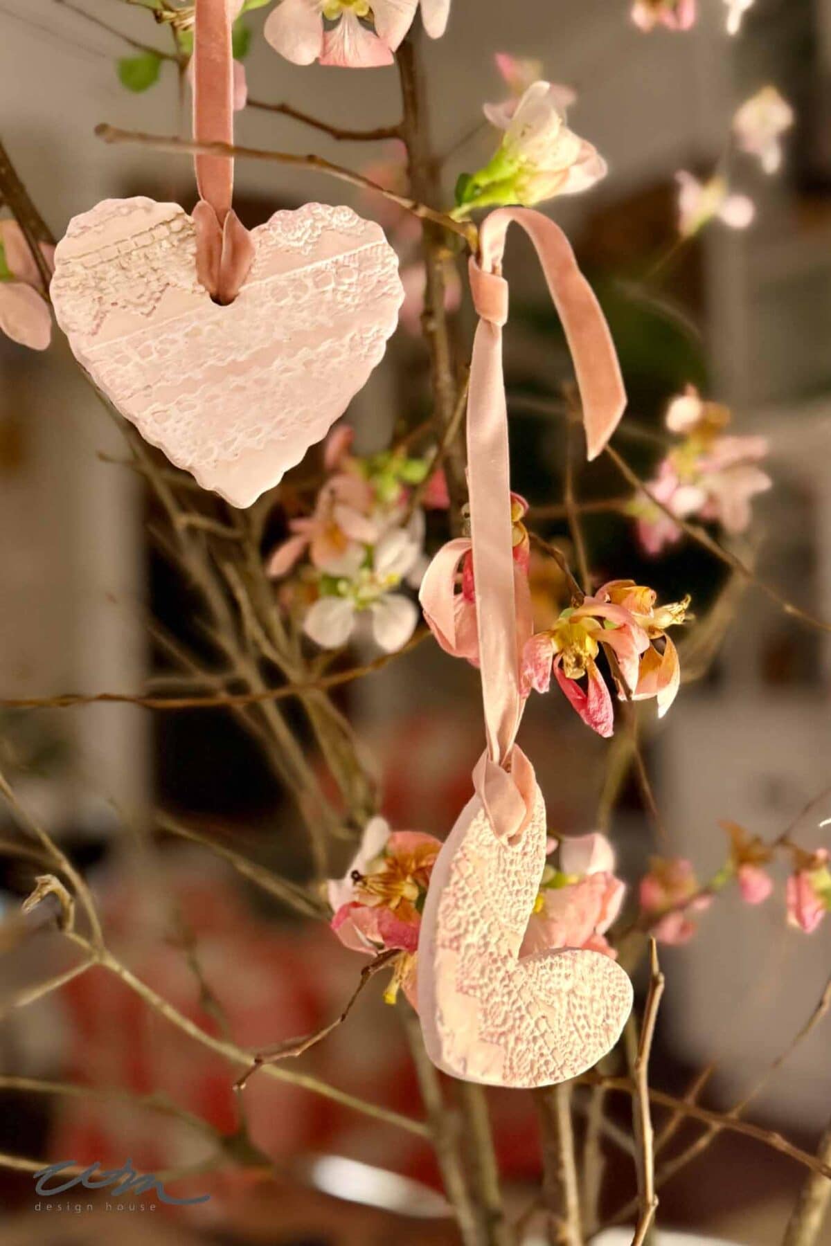 Two lace hearts, crafted with intricate patterns, hang from pink ribbons on a flowering branch adorned with delicate blossoms. The softly blurred background creates a warm, cozy atmosphere.