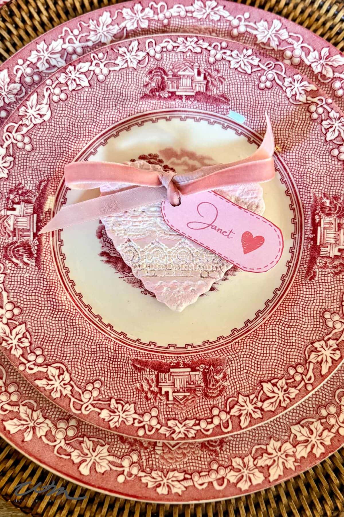 Pink and white decorative plate with a heart-shaped cookie, topped with lace icing in delicate Lace Hearts patterns and a pink ribbon holding a name tag labeled "Janet" with a heart, all set on a woven placemat.