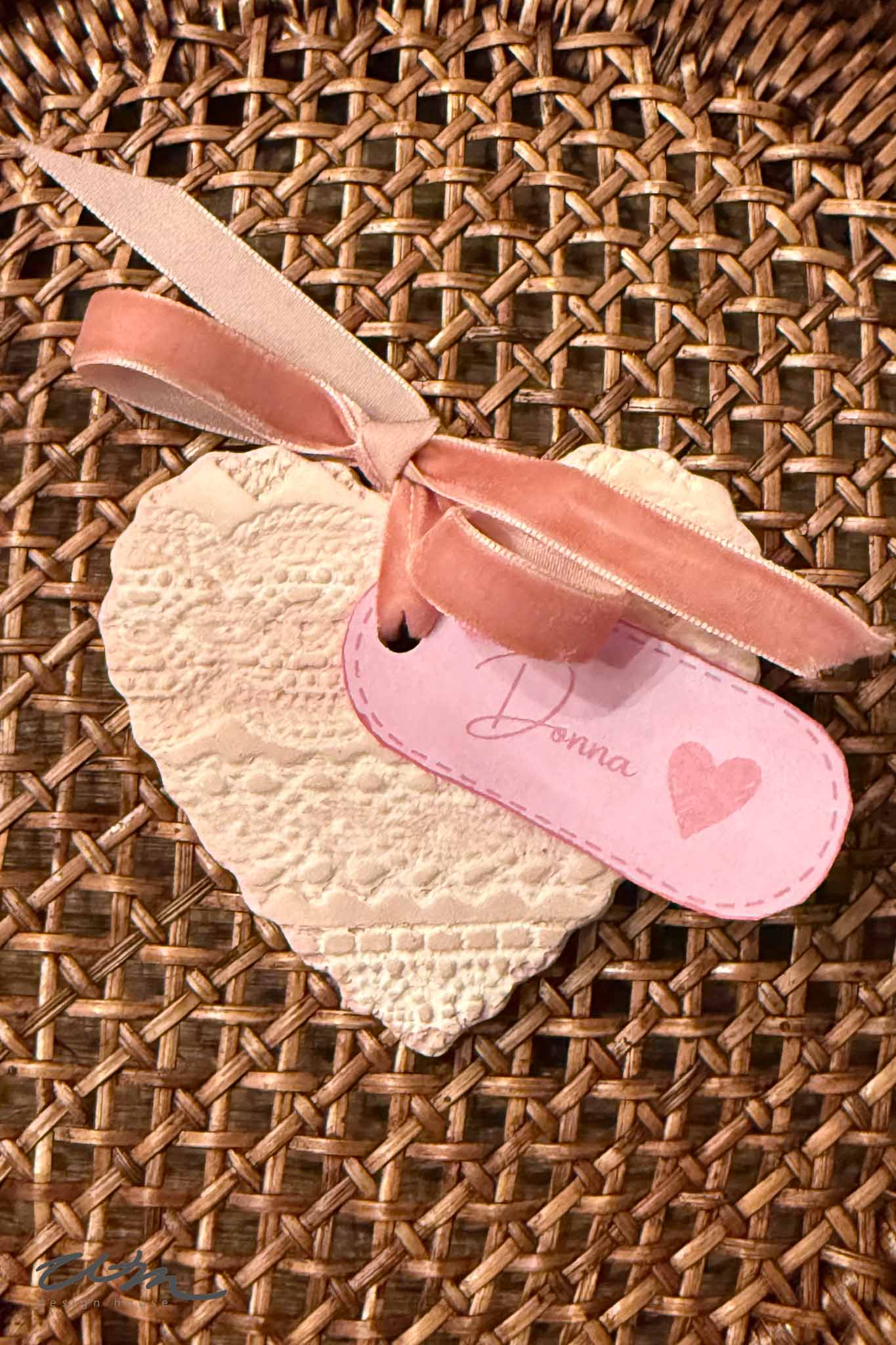 A textured heart-shaped ornament from the Lace Hearts collection, adorned with a pink ribbon and a "Donna" tag with a heart, rests atop a woven wicker surface.