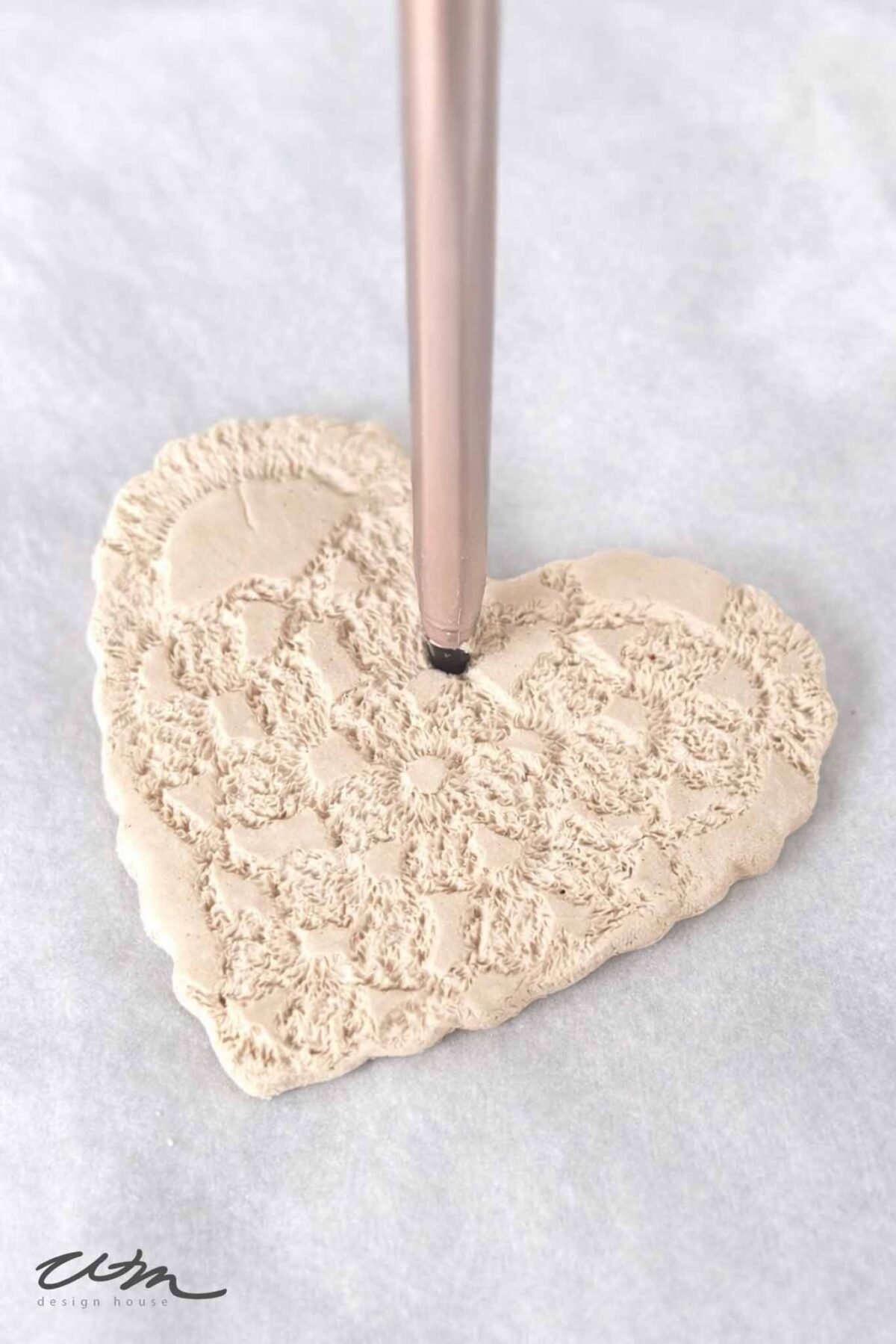 A pen is used to make a hole in the top of a heart-shaped piece of textured clay, perfect for creating delicate Lace Hearts, resting on a white surface.