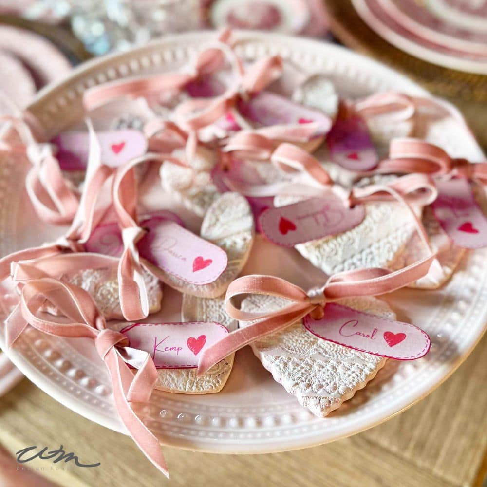 A plate of heart-shaped cookies decorated with lace, each tied with a pink ribbon and a name tag featuring a red heart, arranged for a special occasion or celebration.