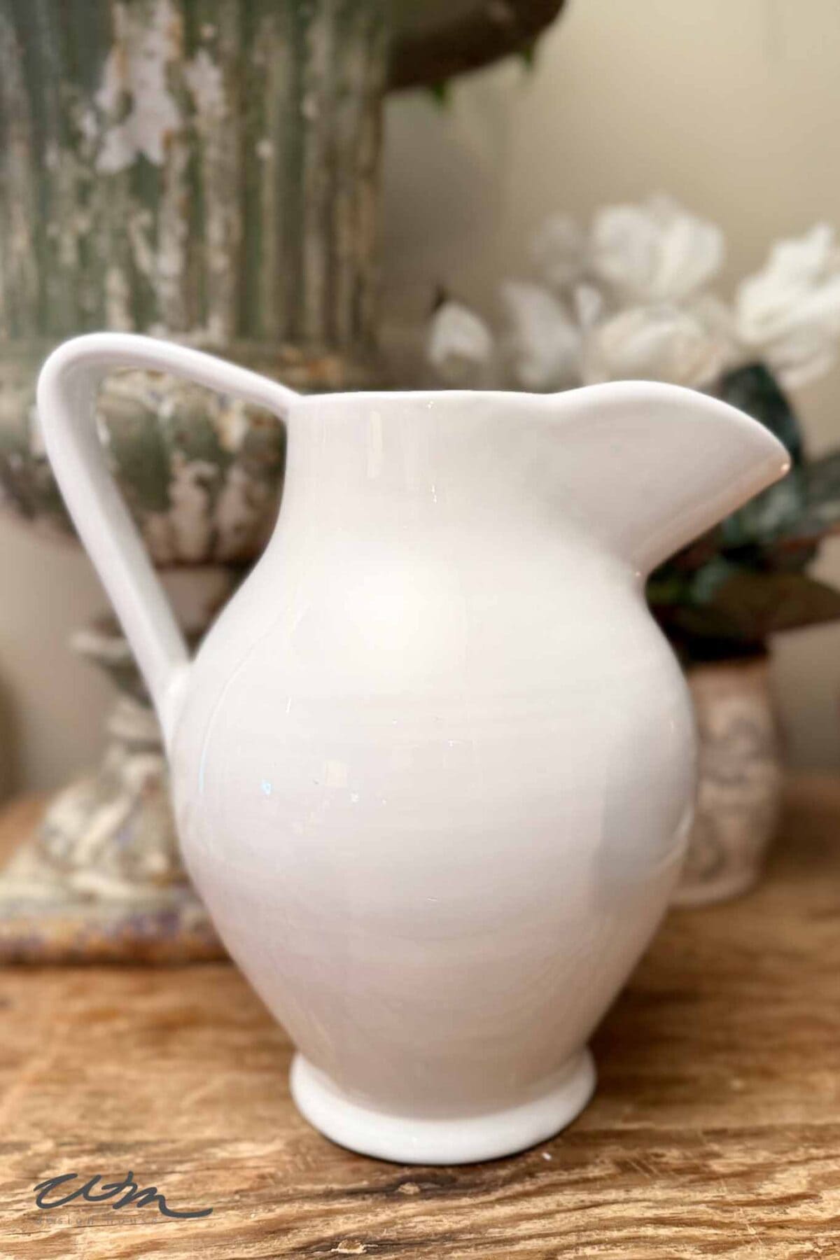 A glossy white ceramic pitcher sits on a wooden surface, bringing vintage charm to the scene, with blurred white flowers and a rustic, weathered vase in the background—perfect inspiration for spring entry table decor.