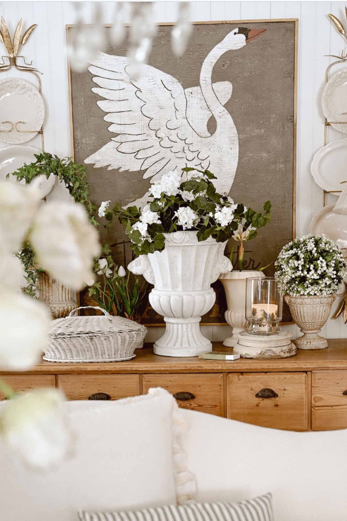 A cozy room with Vintage Charm features a wooden dresser topped with white flower arrangements, a glass candle holder, and wicker trays. A large swan painting and decorative plates add interest—perfect for Spring Entry Table Decor.