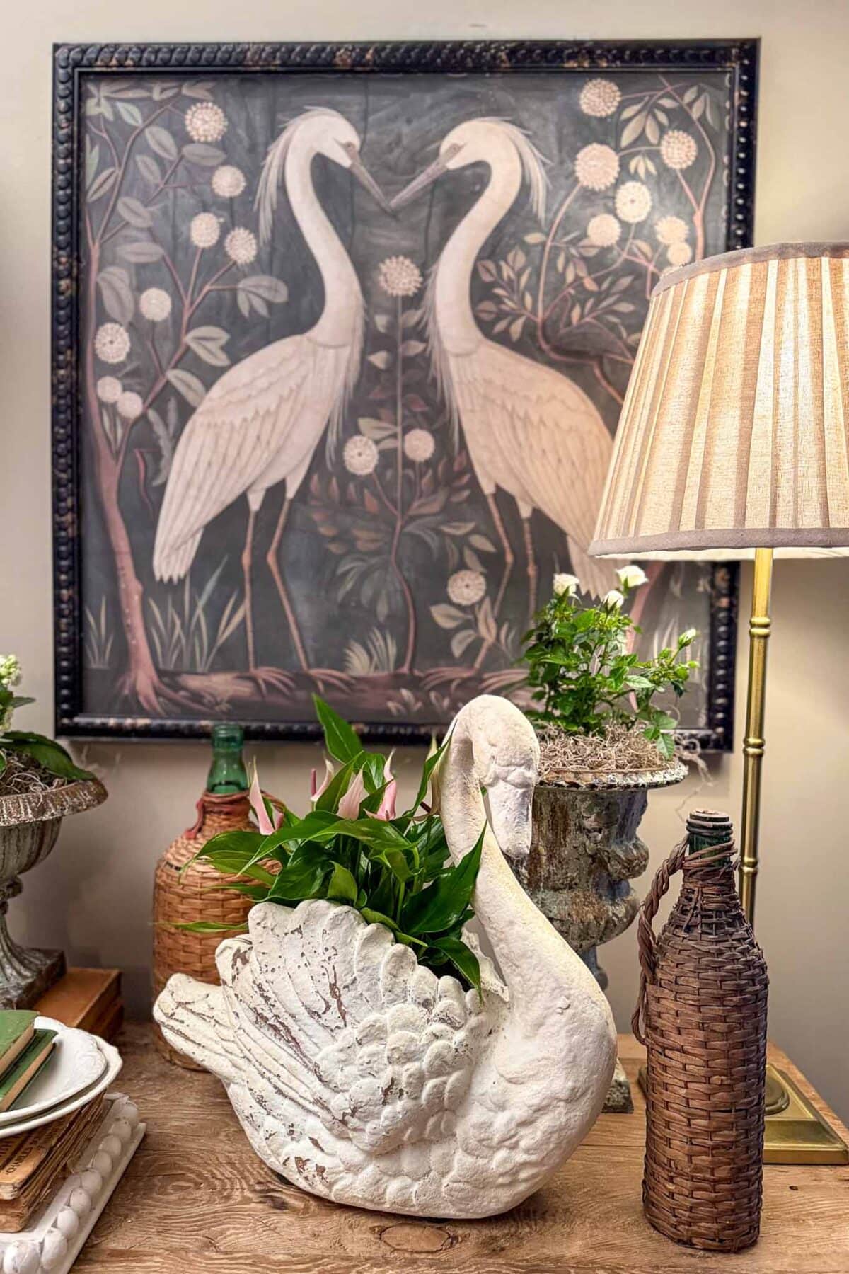 Brimming with vintage charm, this spring entry table decor features a white swan planter with lush greenery, vintage bottles, and a lamp. Behind it, artwork of two white cranes among flowers and foliage completes the elegant scene.