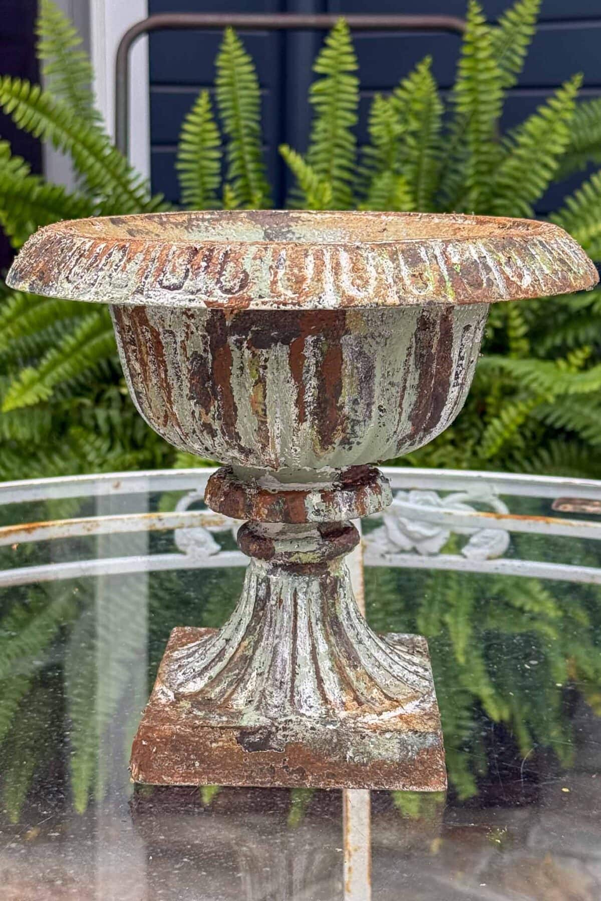 A weathered, vintage metal urn with peeling paint sits on a glass table outdoors. Lush green ferns are in the background, giving a natural, garden setting.